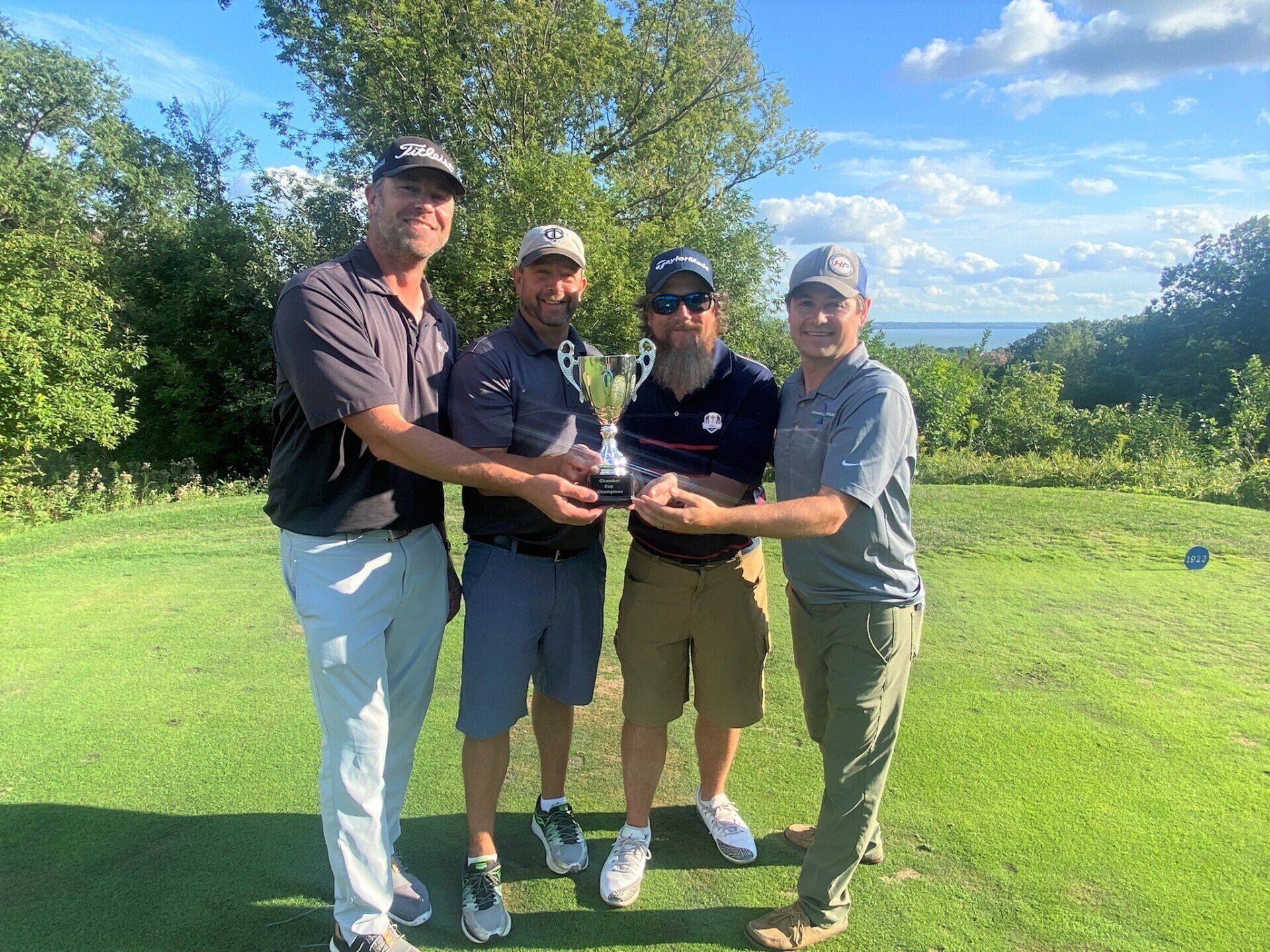 Four golfers smiling while holding a trophy on a grassy course with a scenic landscape background.