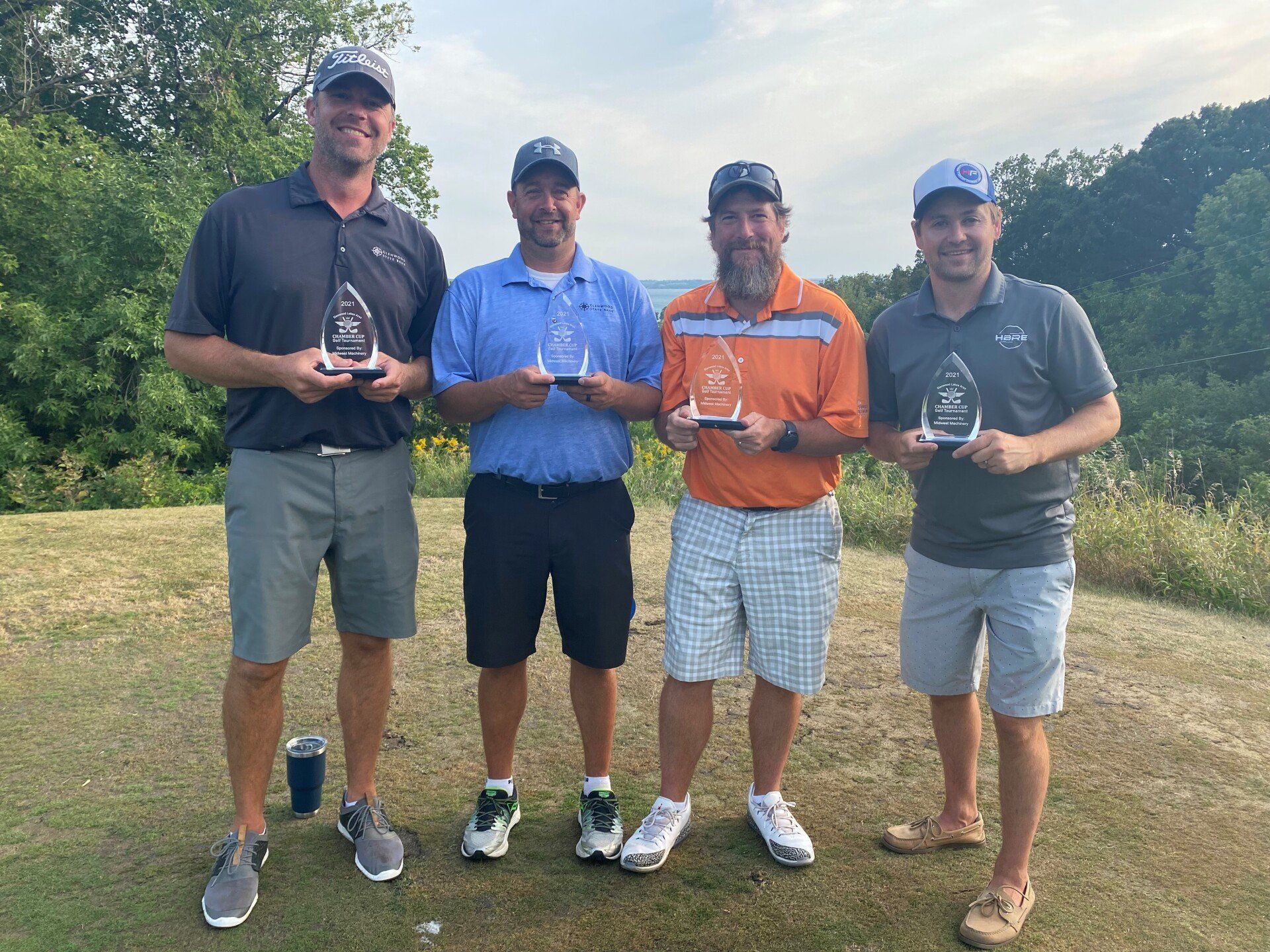Four men in hats and golf attire pose outdoors holding trophies, with a scenic, grassy landscape in the background.