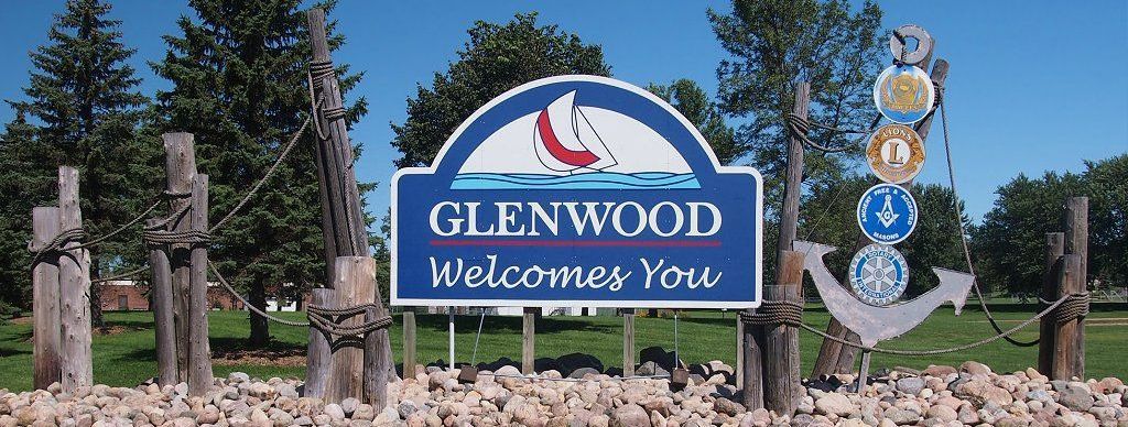 A blue and white welcome sign for Glenwood, featuring a sailboat logo, an anchor, and wooden posts in a grassy setting.