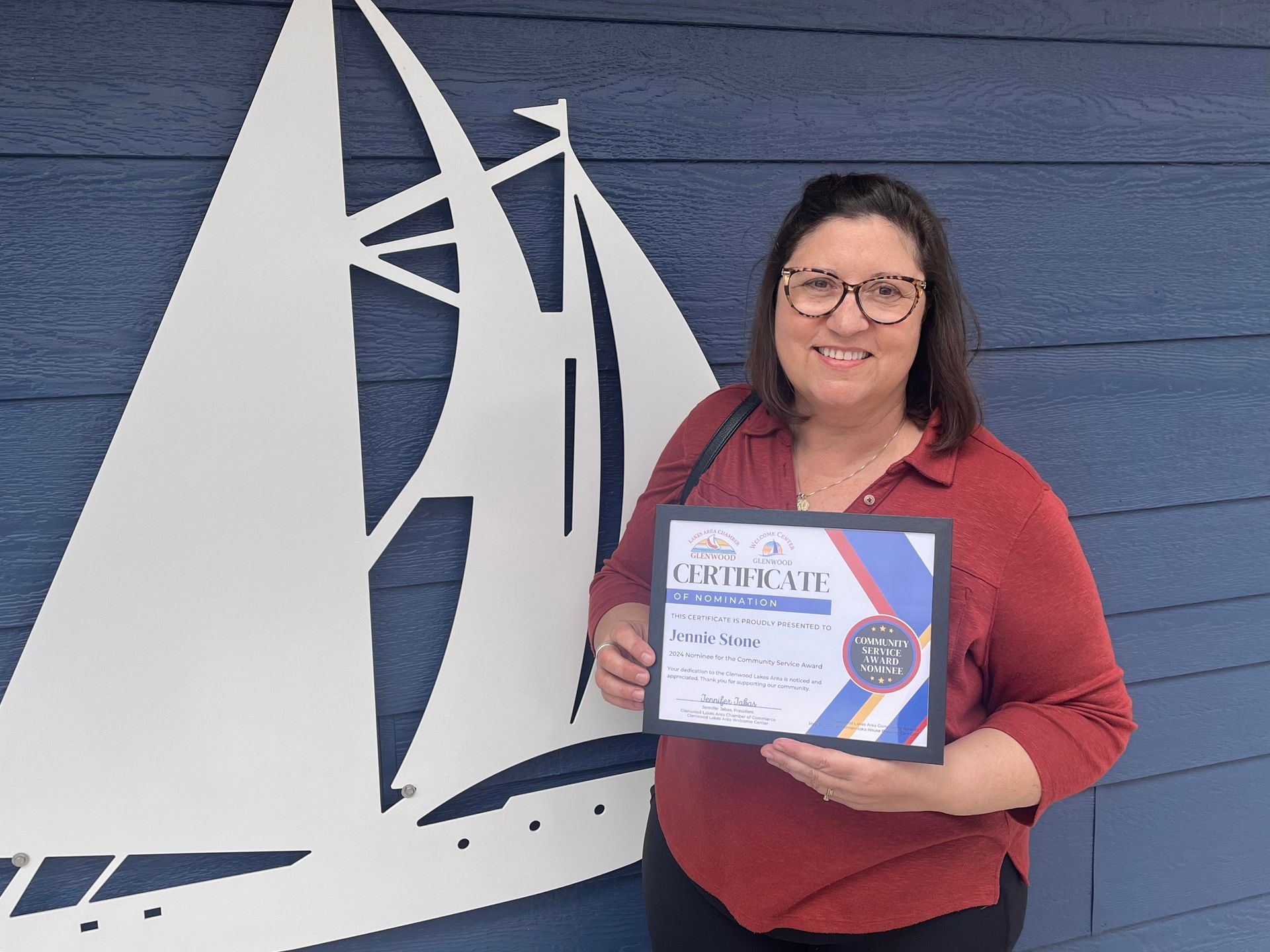 A smiling person wearing glasses and a red shirt holds a certificate in front of a white sailboat wall art.