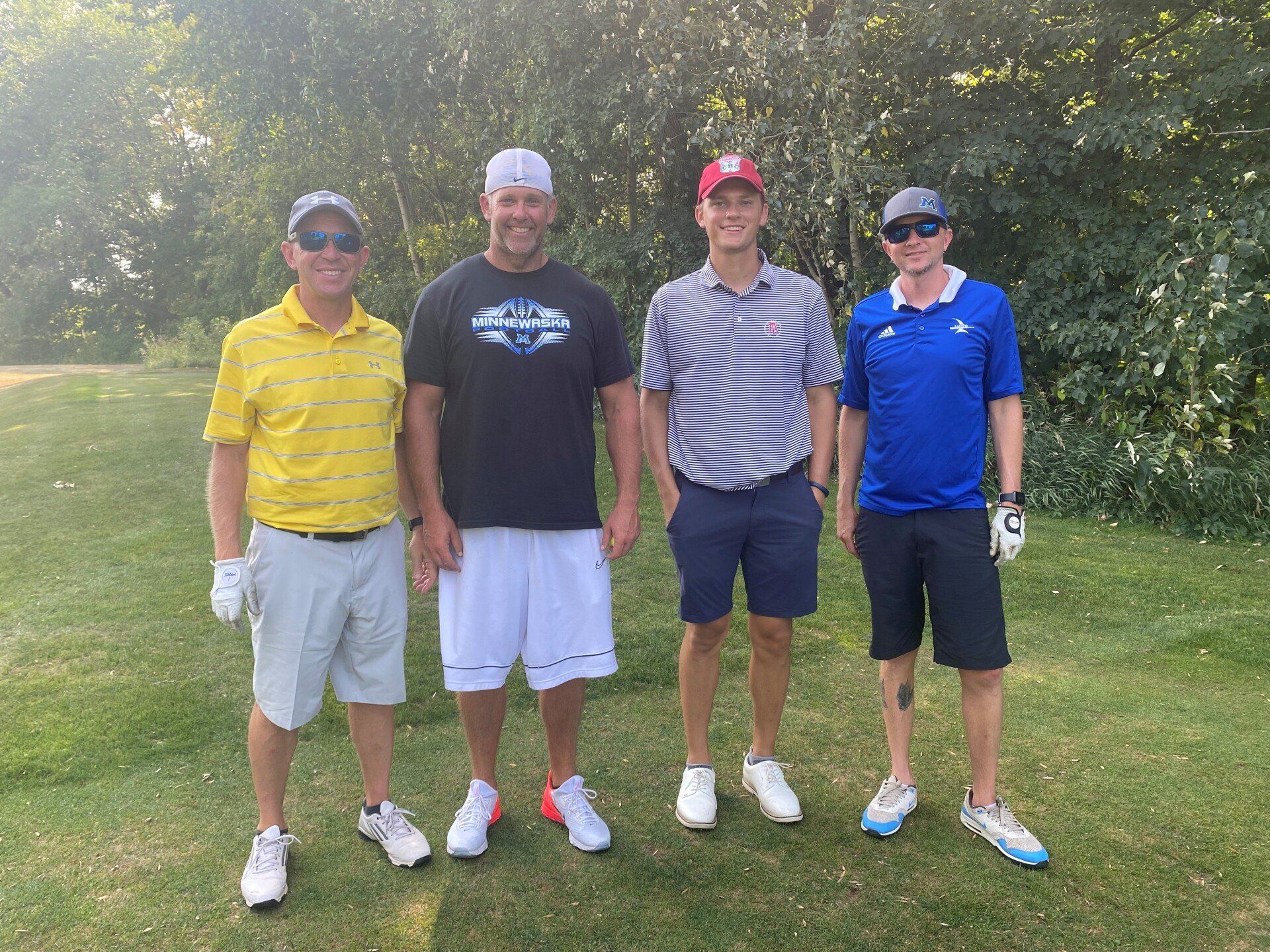 Four people in golf attire stand together outdoors on a grassy course, smiling at the camera.