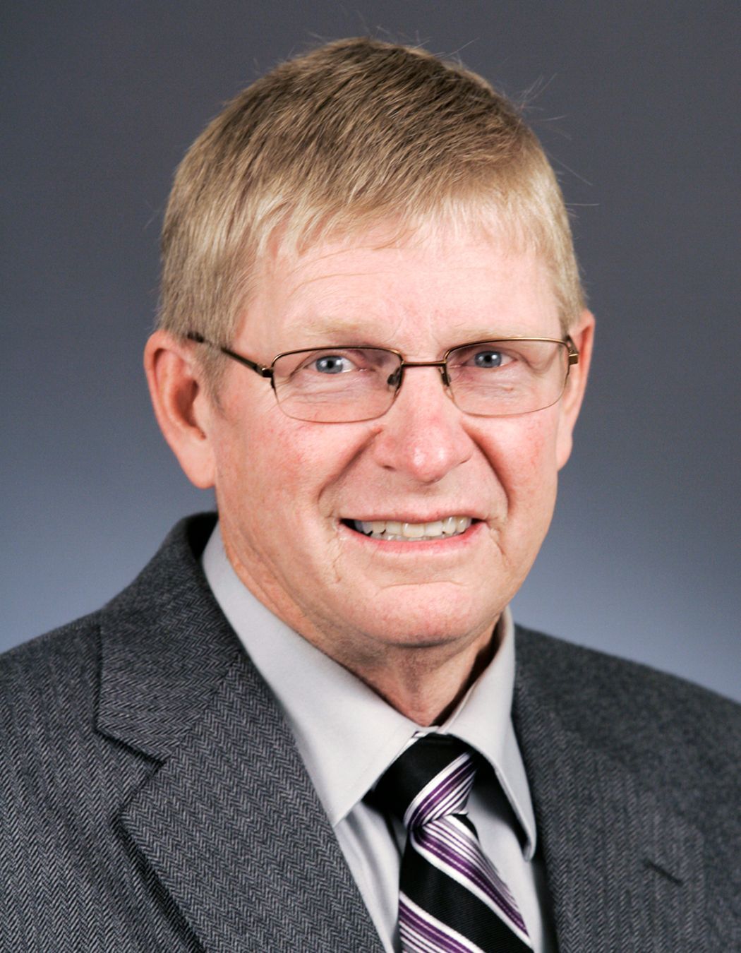 A person wearing glasses, a grey suit jacket, and a striped tie against a plain grey background.