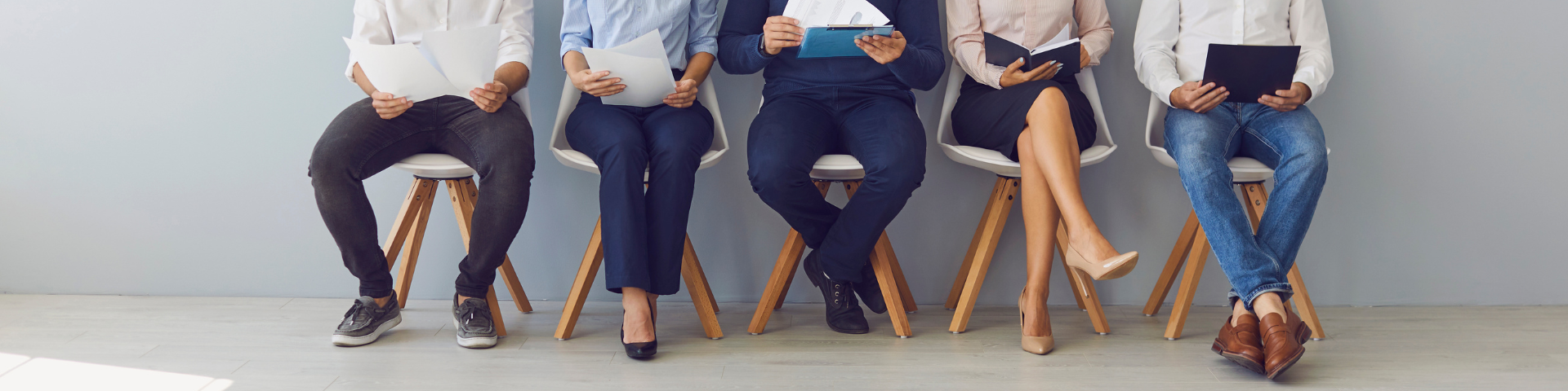 Five people in business casual attire sit in a row on chairs against a plain wall, holding folders or tablets.