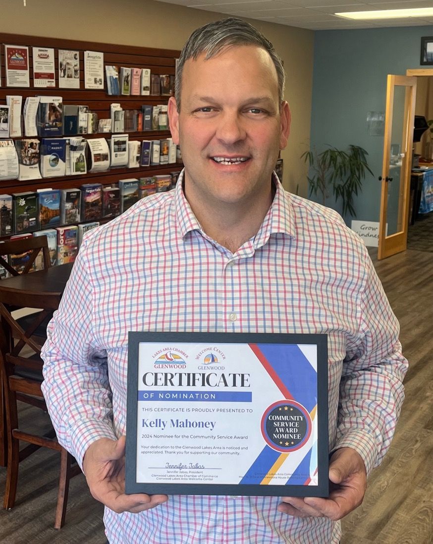 A person in a checkered button-down shirt smiling while holding a framed certificate in an office with shelves of brochures.