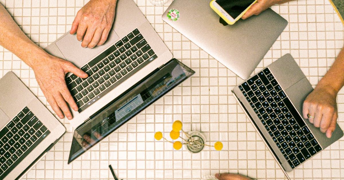 A high-angle view of hands working on three laptops on a white tiled table, with a small yellow decorative object nearby.