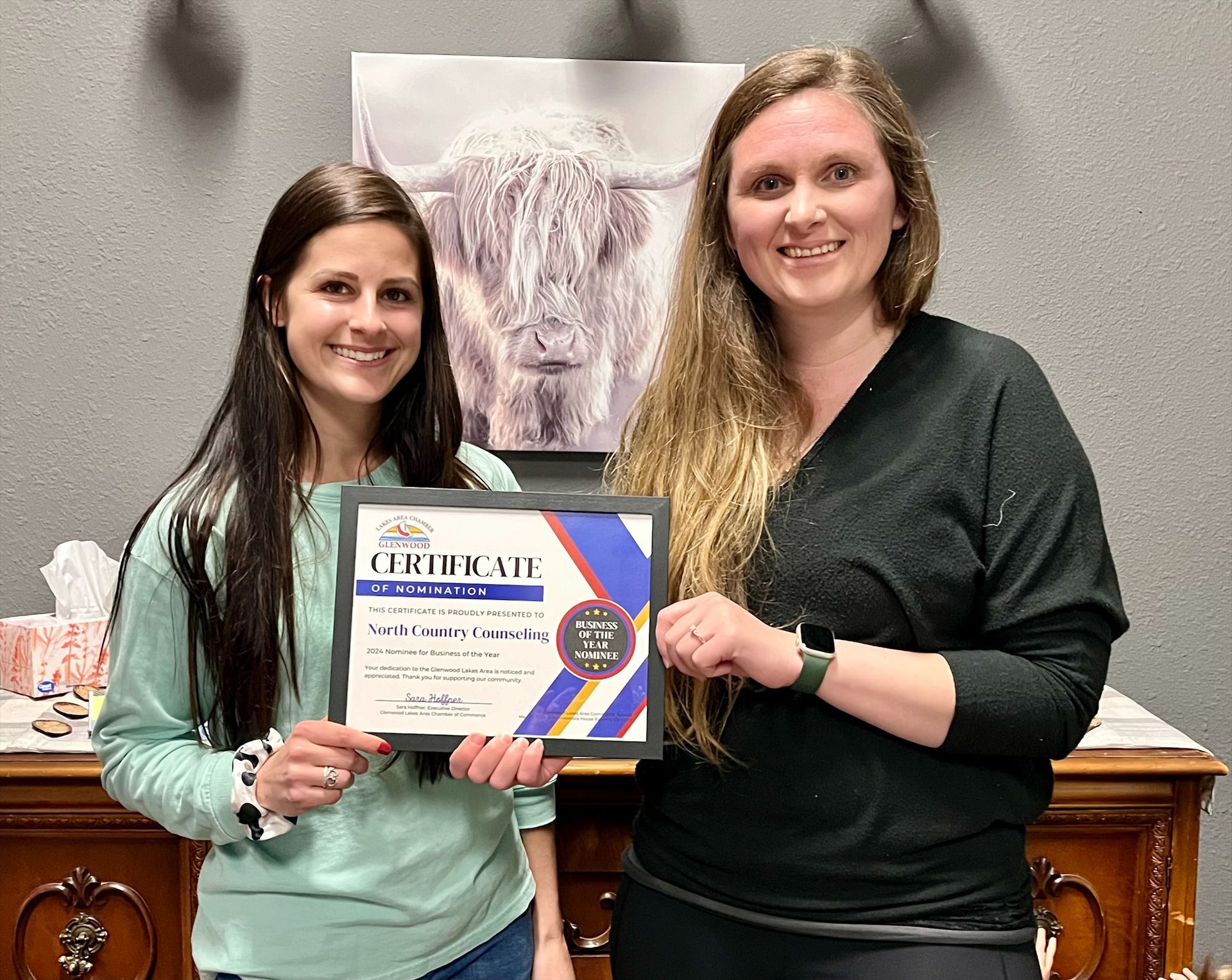 Two smiling individuals hold a framed certificate in front of a wall featuring a large print of a longhorn cow.