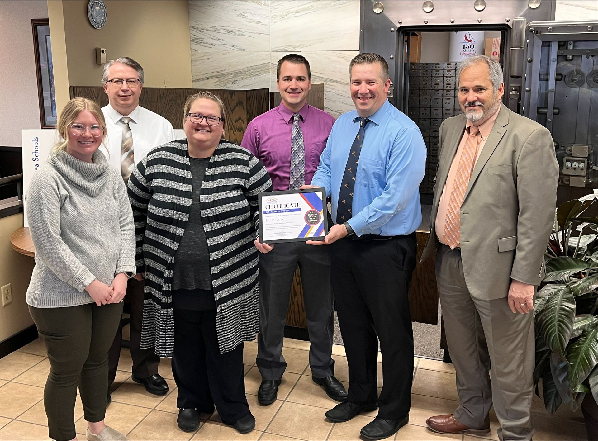 Six people stand in an office posing for a photo while two of them hold a framed certificate in front of a vault door.