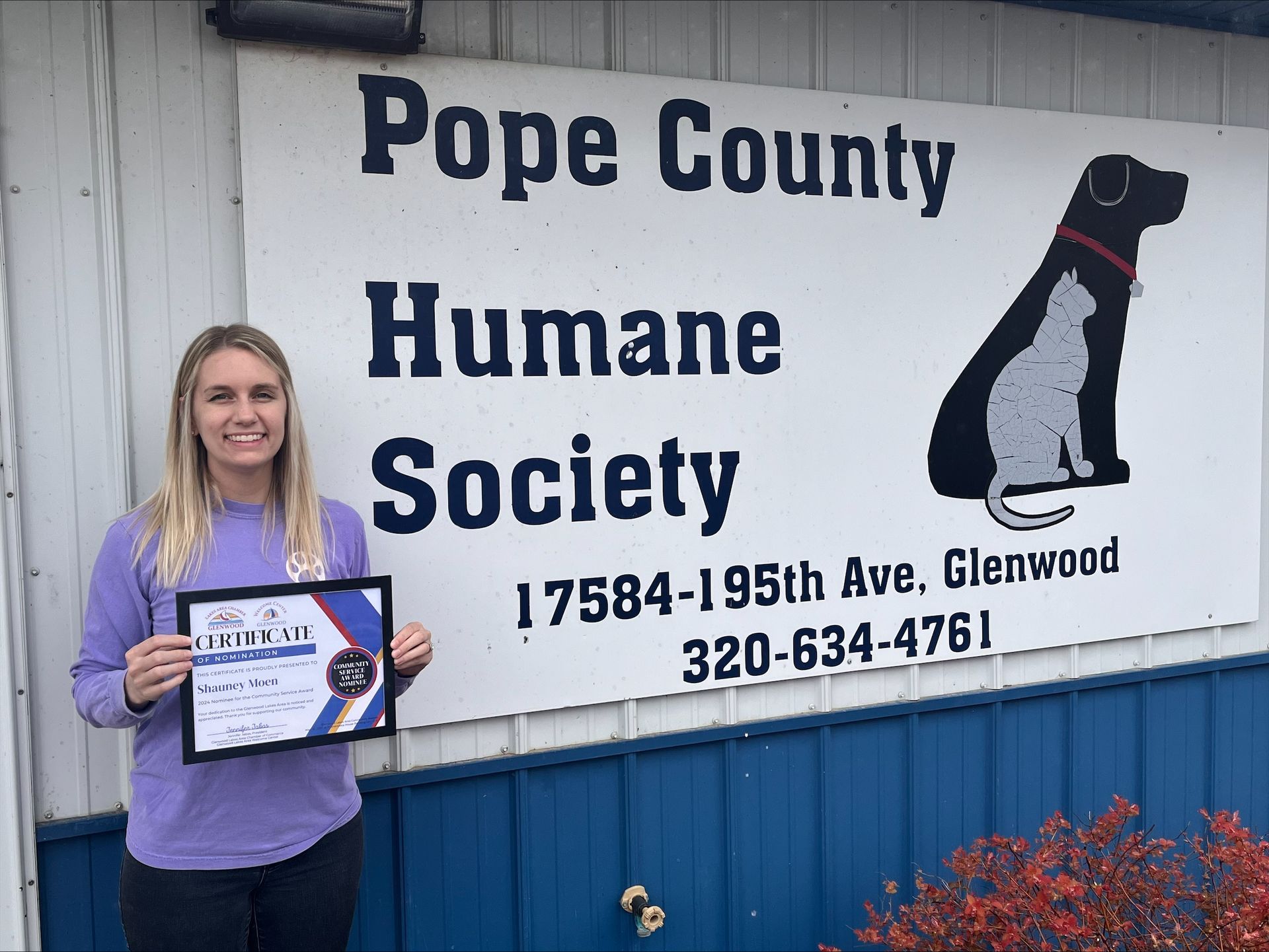 A smiling person stands outside the Pope County Humane Society, holding a certificate in front of a white building sign.