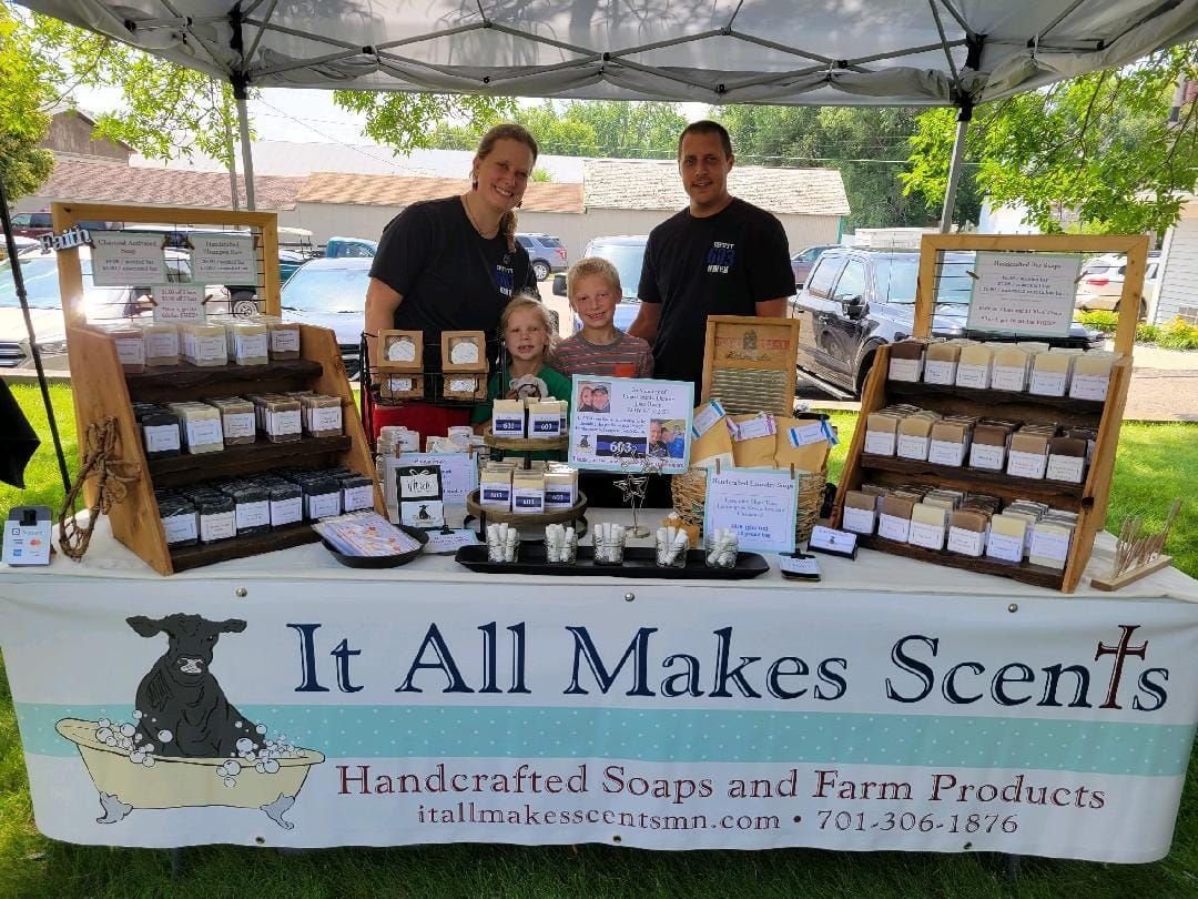 A family stands behind a booth displaying handmade soaps at an outdoor market with a banner reading,