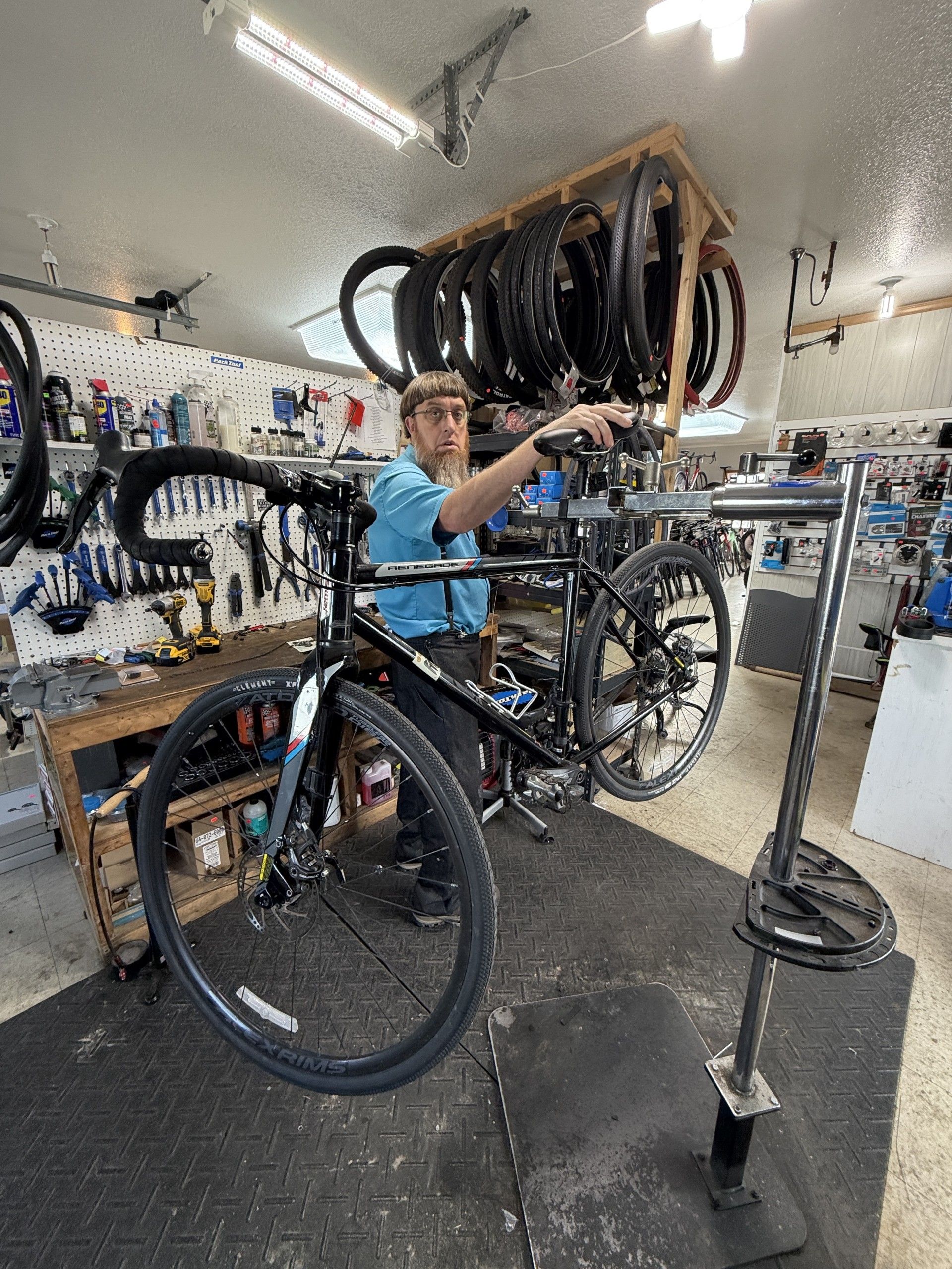 A technician in a blue shirt works on a black bicycle suspended in a repair stand inside a bike shop.