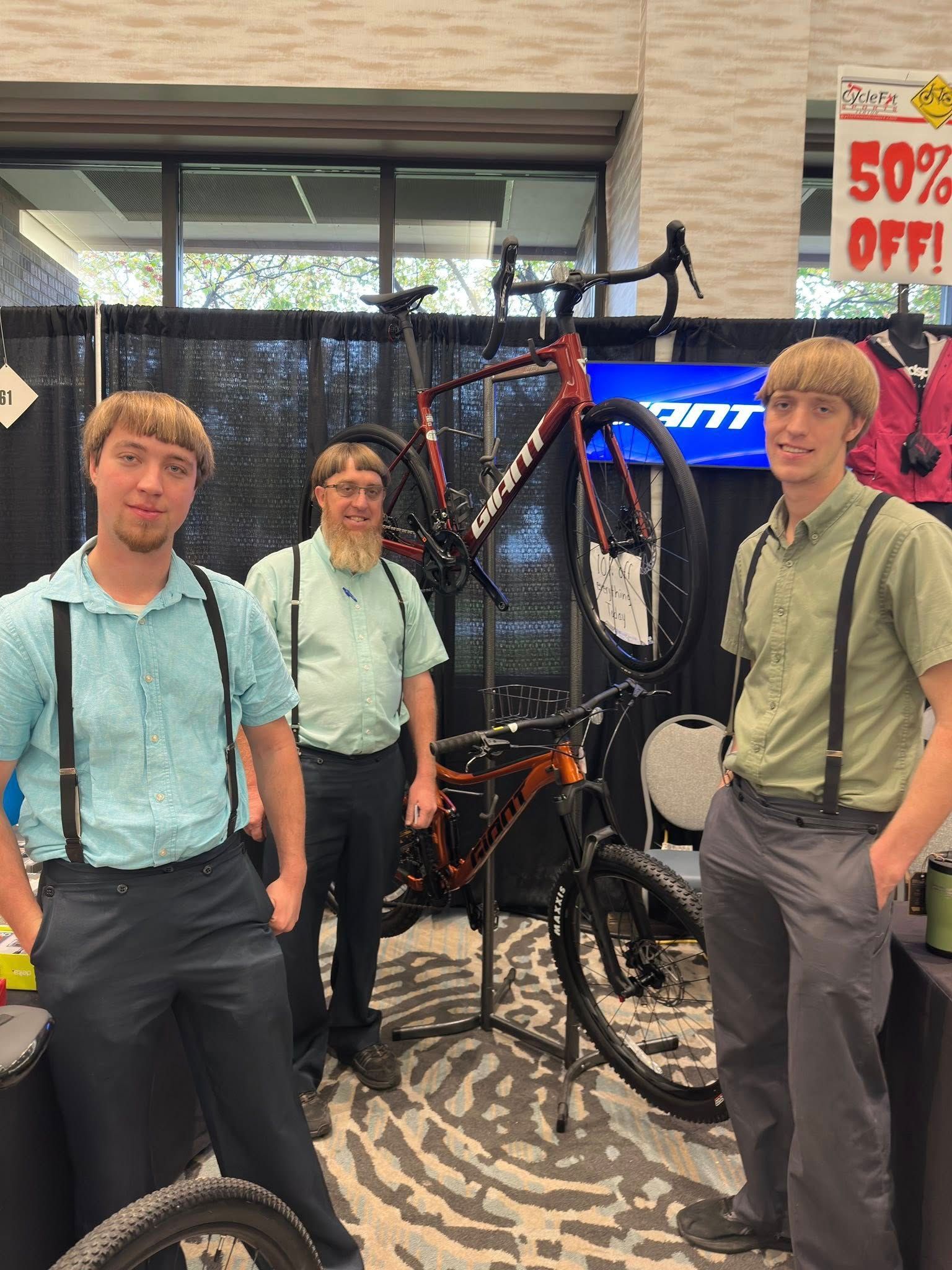 Three people stand in front of two bicycles displayed at a booth with a 50% off sign in the background.