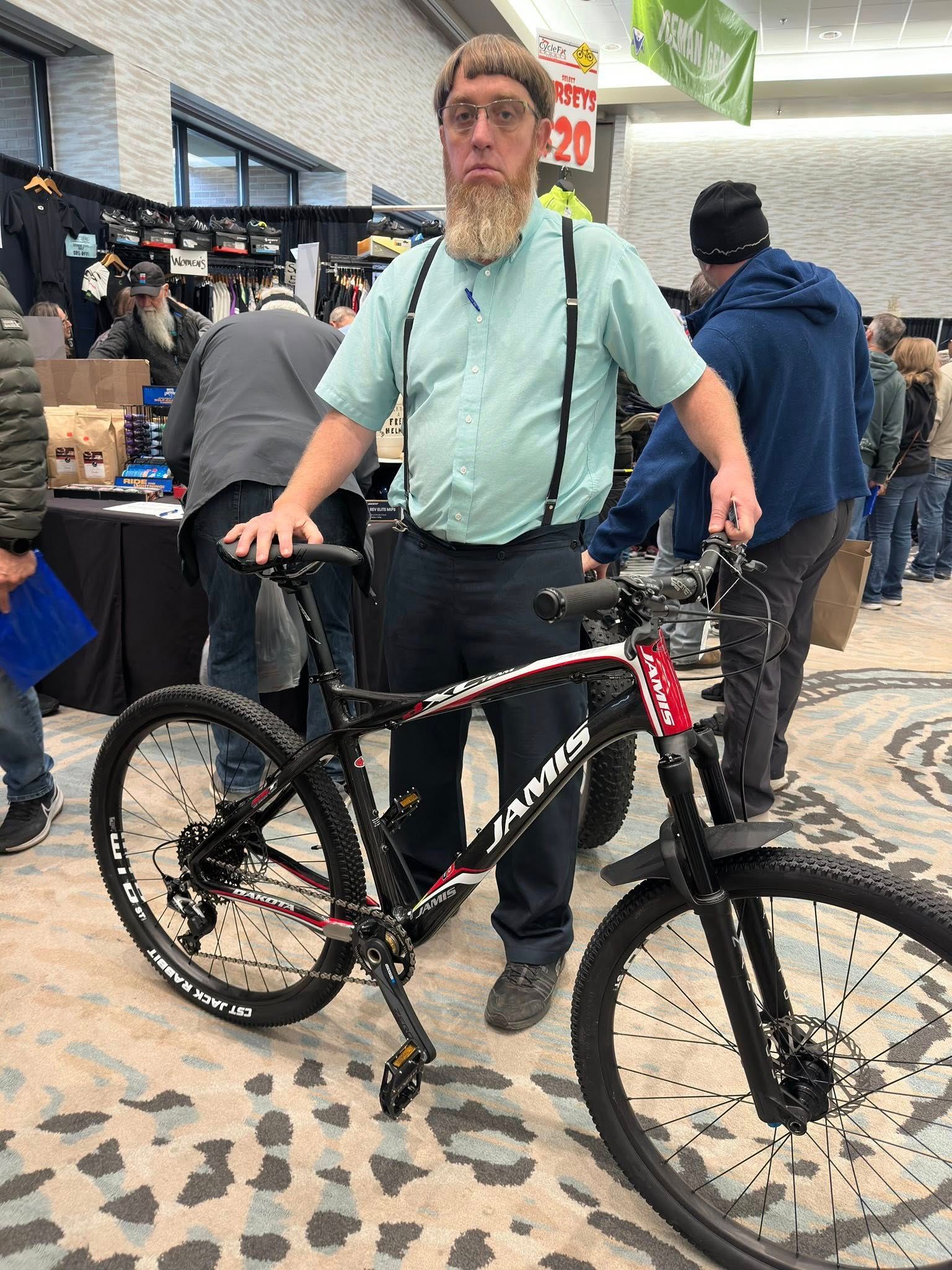 A person with a beard and suspenders stands indoors holding a black mountain bike with red accents.