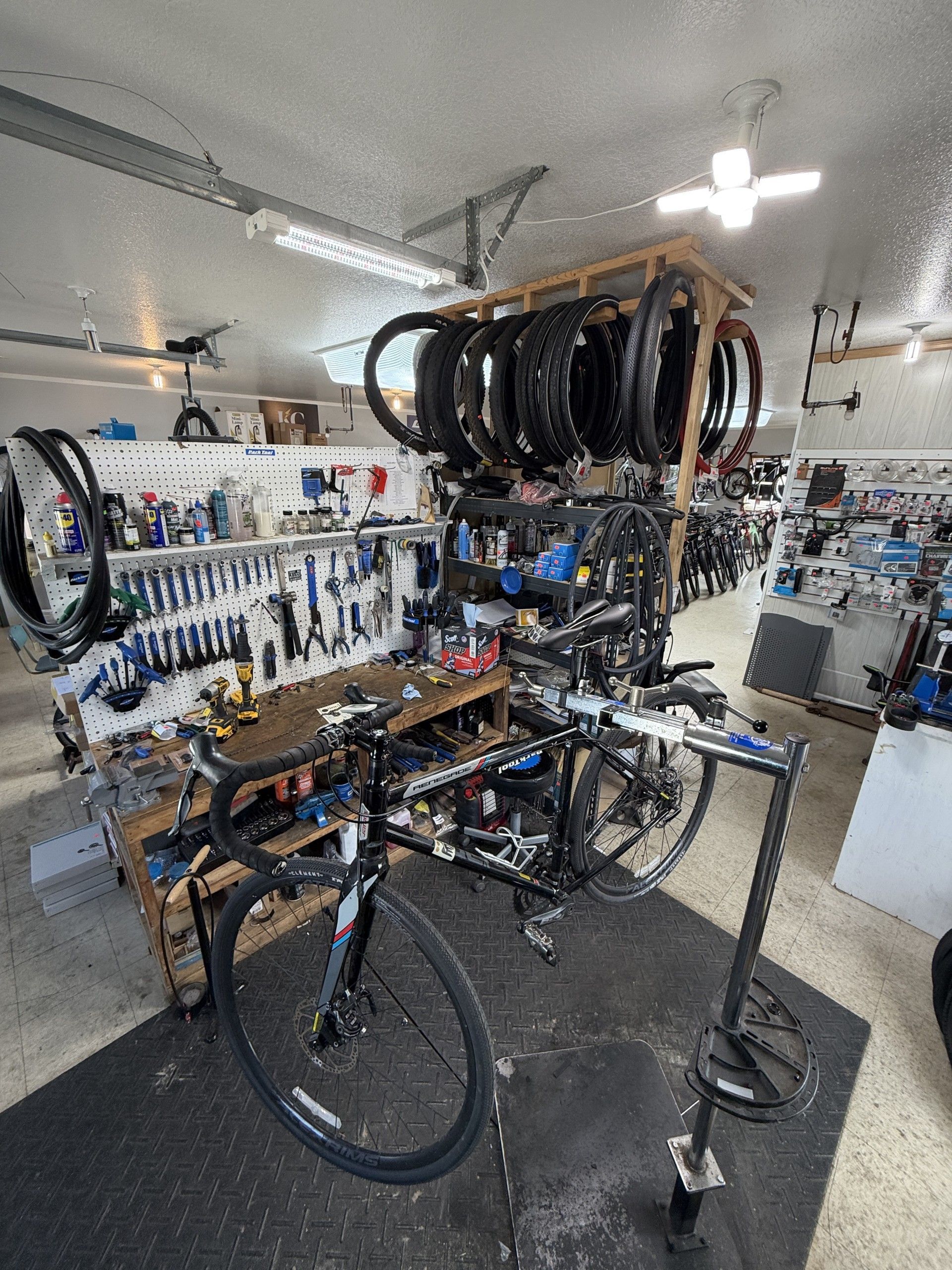 A black road bike on a repair stand in a bike shop, with a workbench and a rack of hanging bicycle wheels in the background.