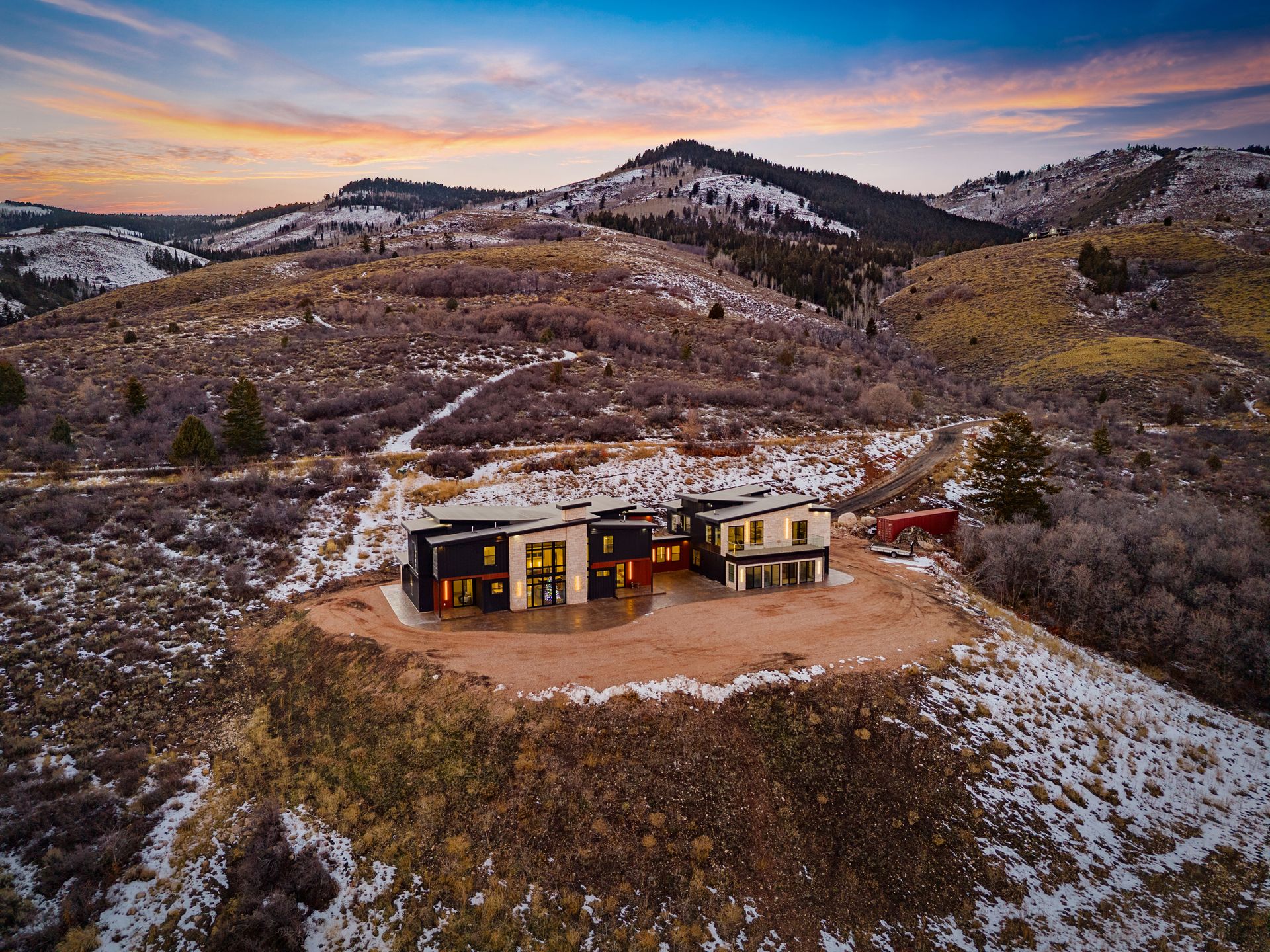 Small modern cabin in snowy hills at sunset, with warm lights and a winding dirt path.