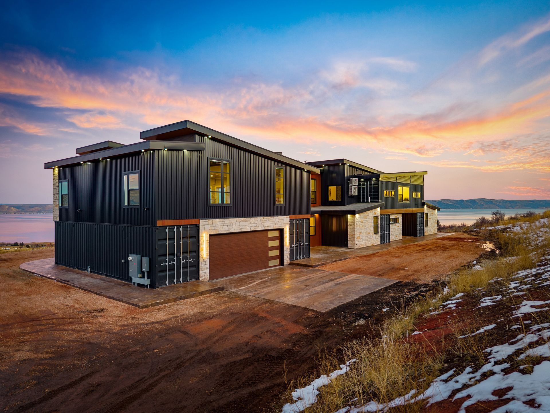 Modern house under construction at sunset, with dark siding, dirt lot, and patches of snow nearby.
