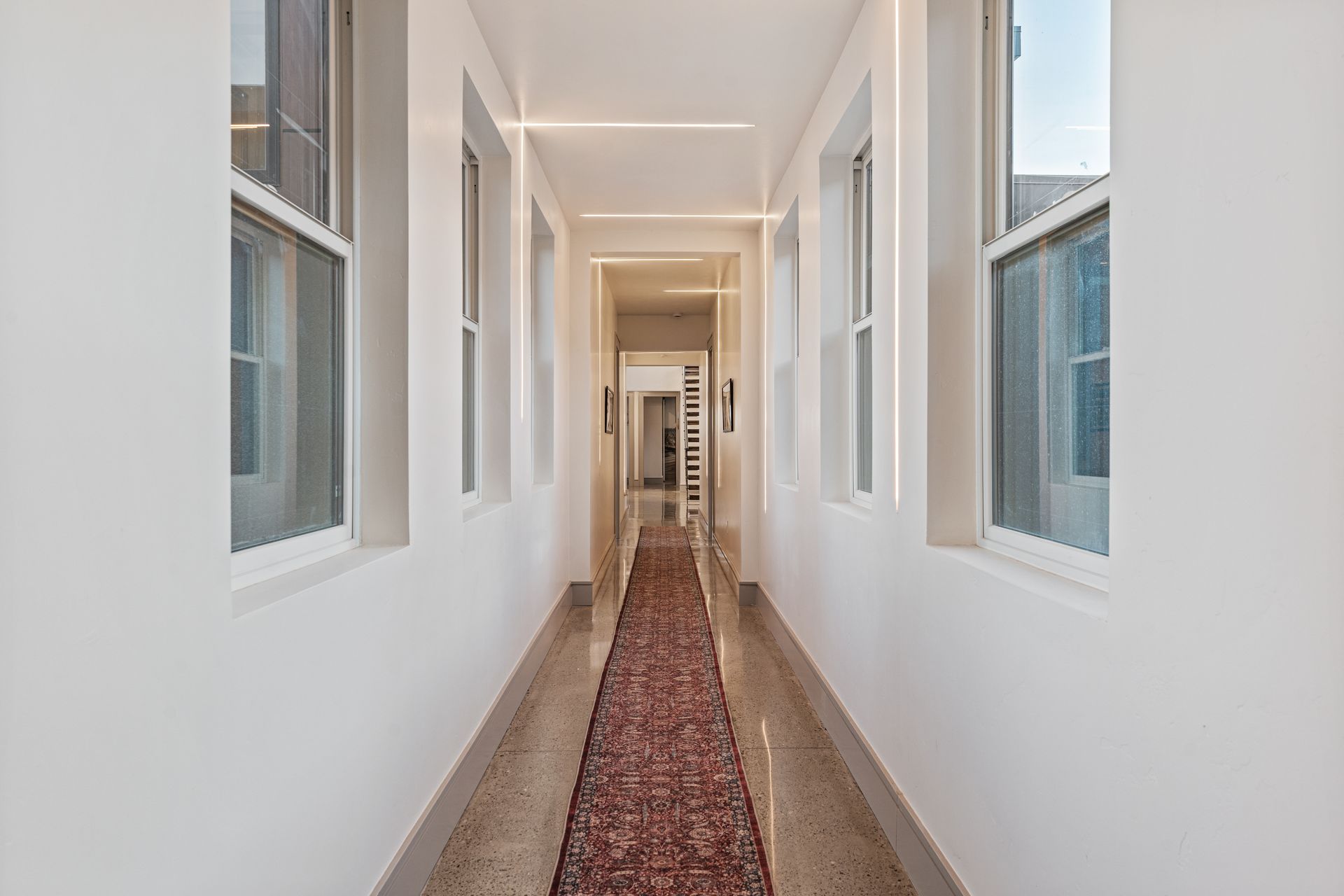 Long, bright hallway with white walls, windows, and a red patterned runner rug