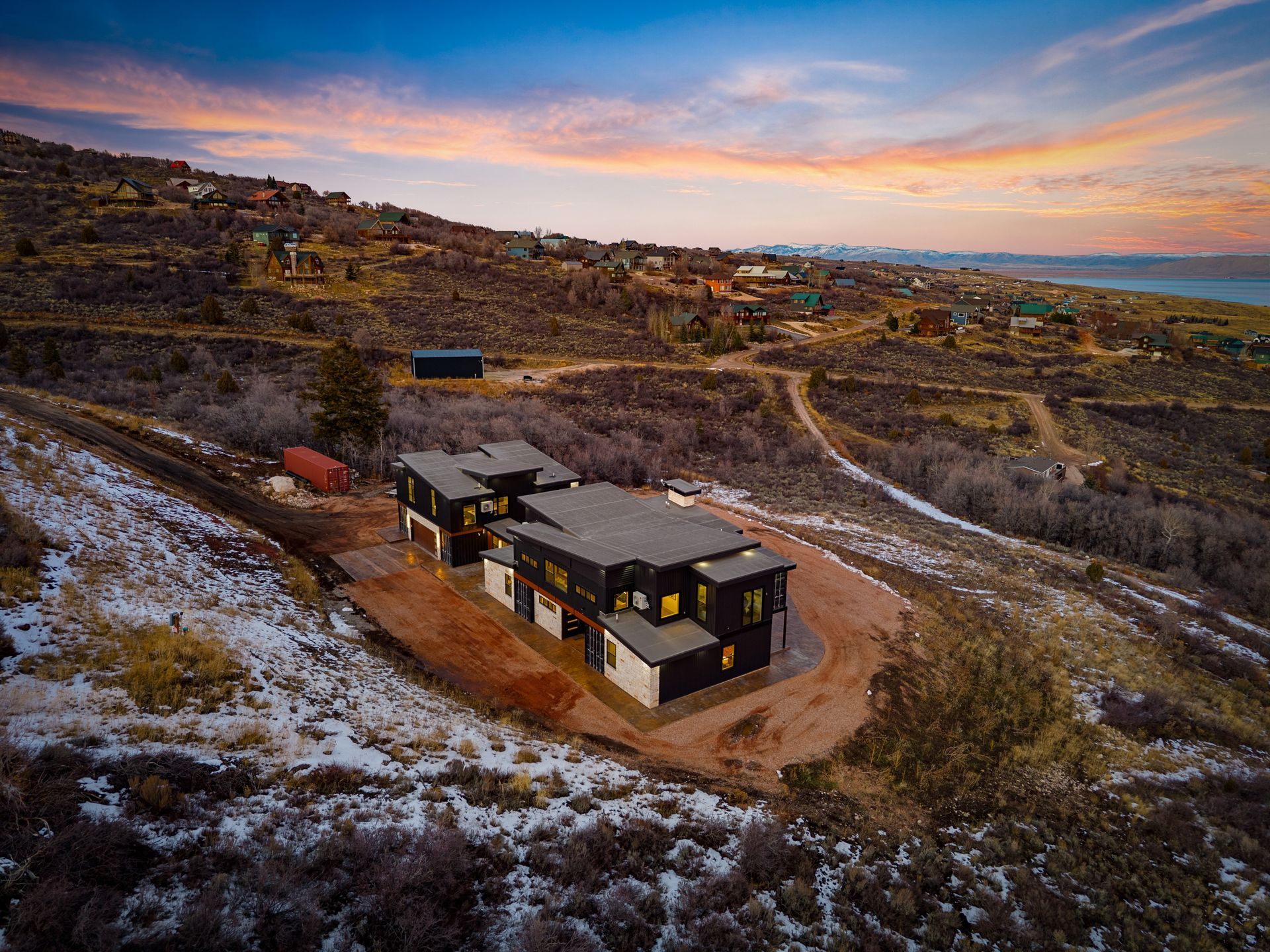 Modern house on a snowy hillside at sunset, with winding dirt paths and distant hills under a colorful sky