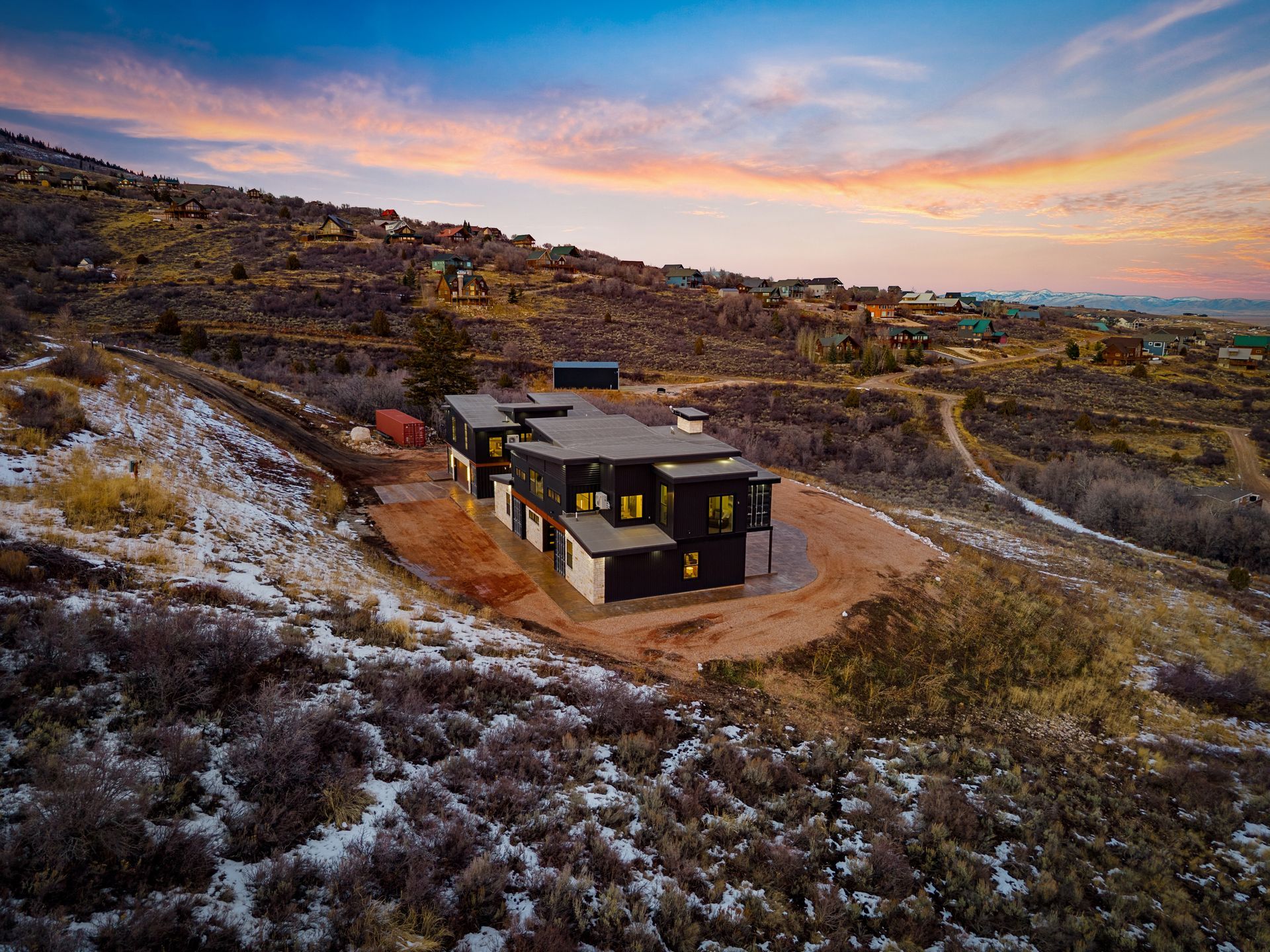 Small modern cabin on a frosty hillside at sunset, with pink clouds over a rugged landscape.