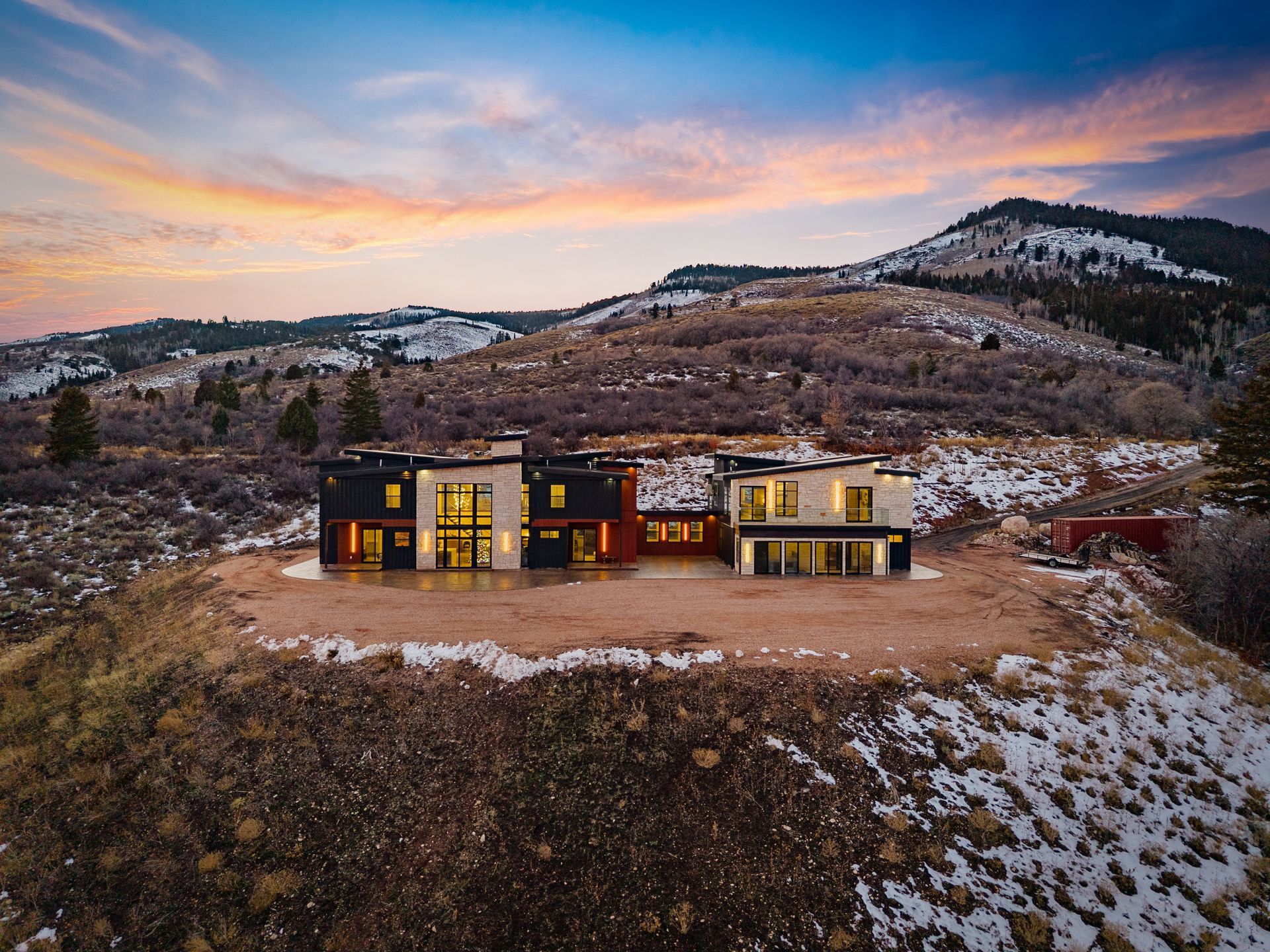 Modern house at dusk on a snowy hillside, with mountains and a pink-orange sky in the background