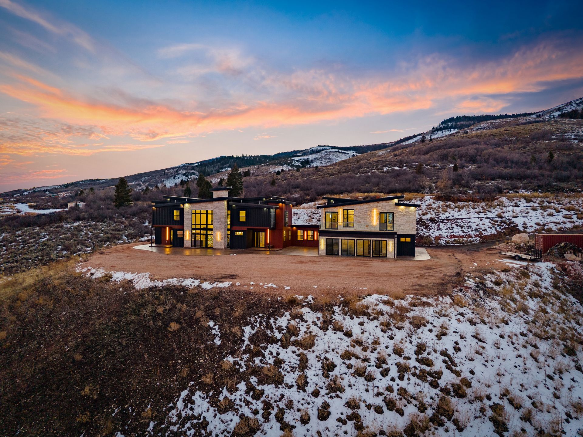 Modern house lit at dusk in a snowy hillside landscape under a pink sunset