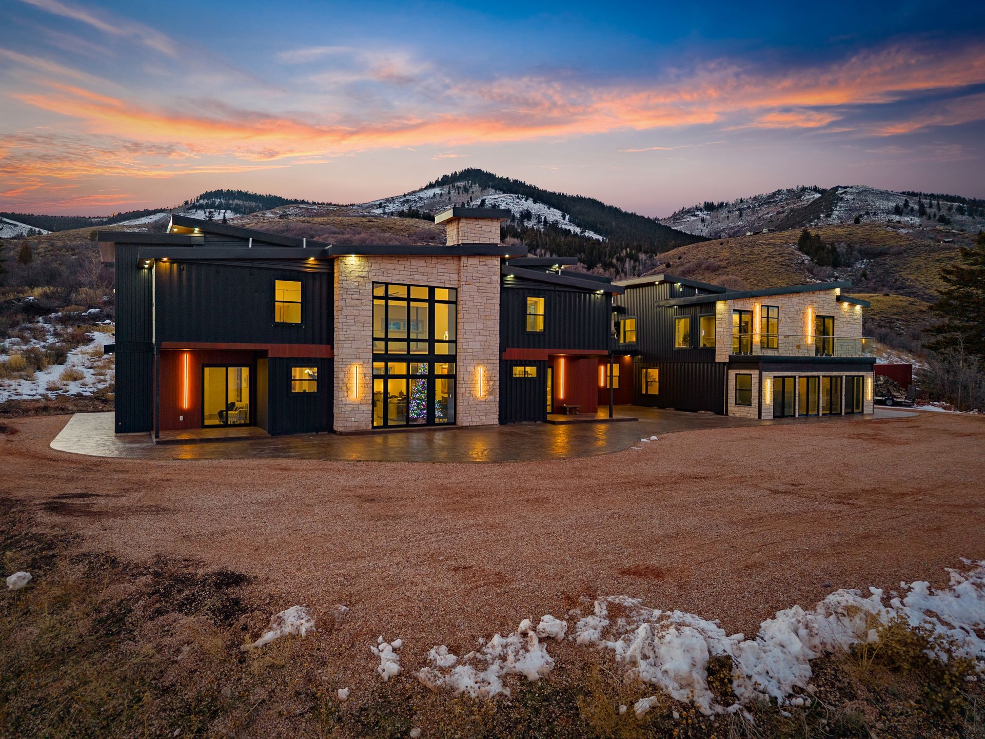 Modern luxury house at sunset with warm lights, dark exterior, and mountain backdrop