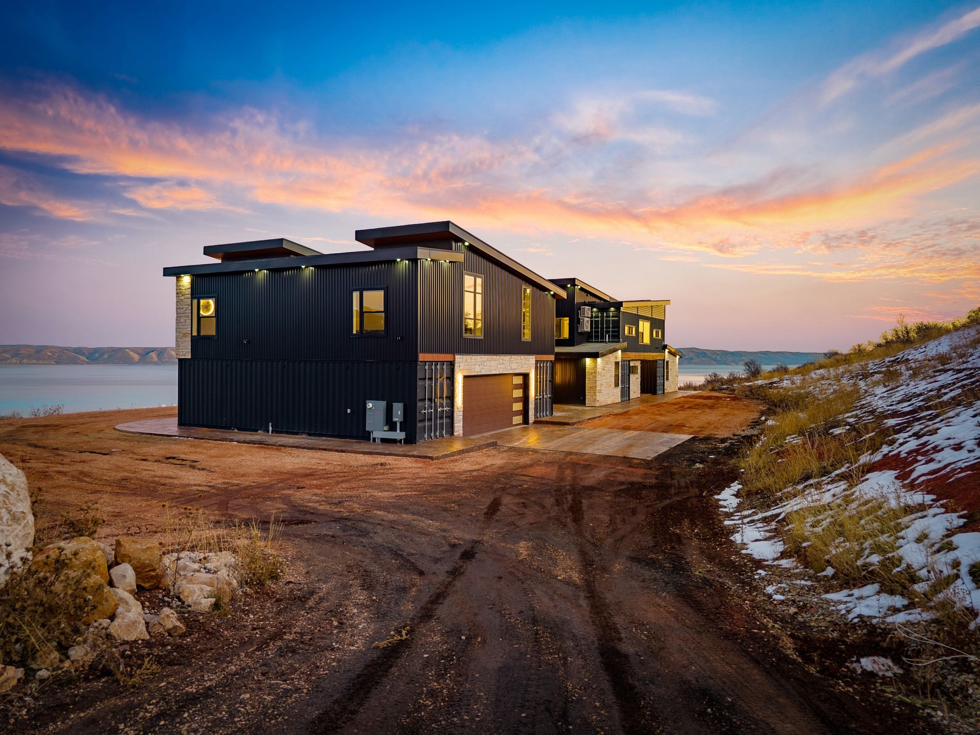 Dark modern houses on a snowy coastal path at sunset, with ocean and colorful sky in the background
