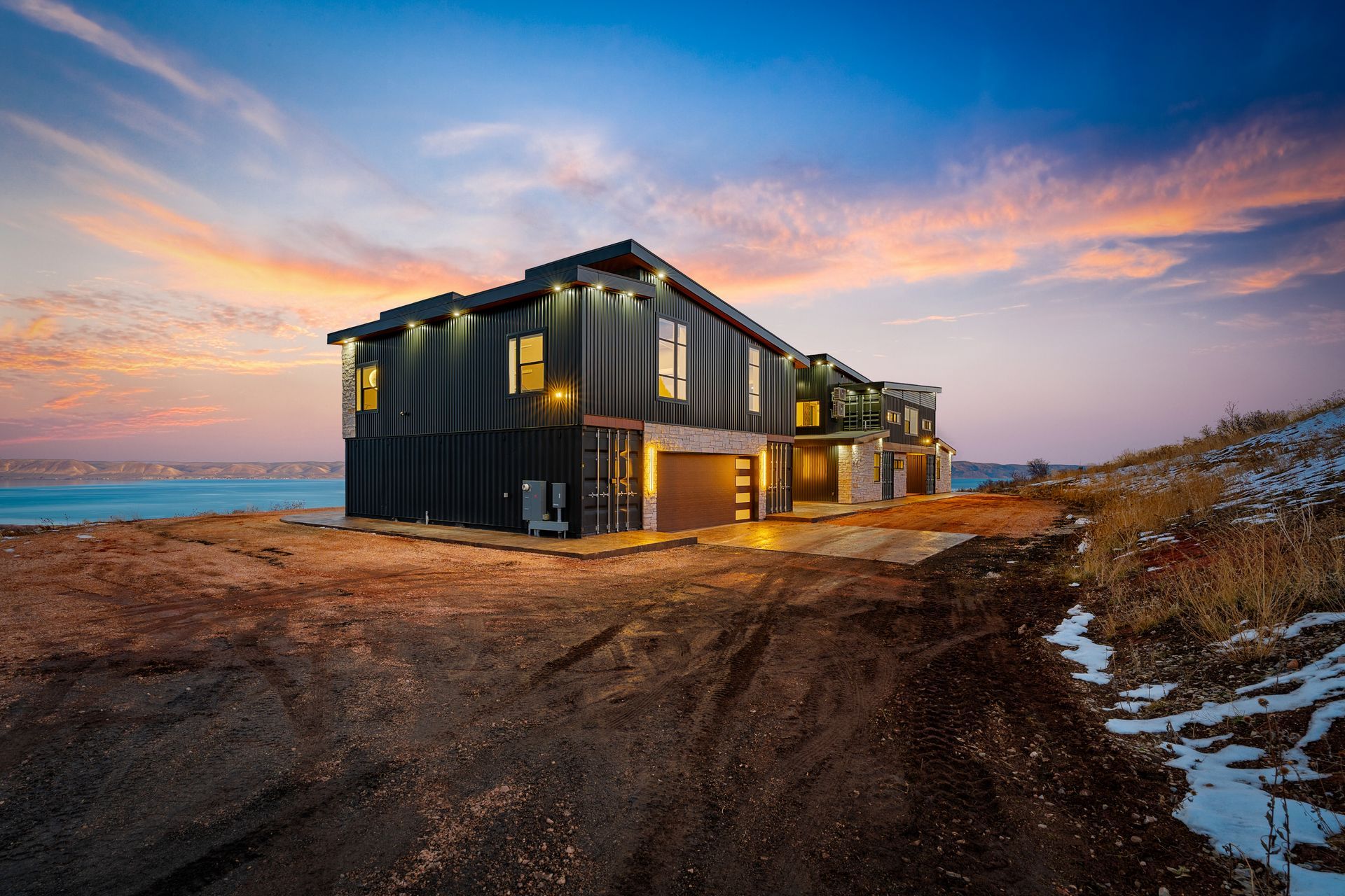 Modern black house on a coastal cliff at sunset with glowing windows and snowy ground