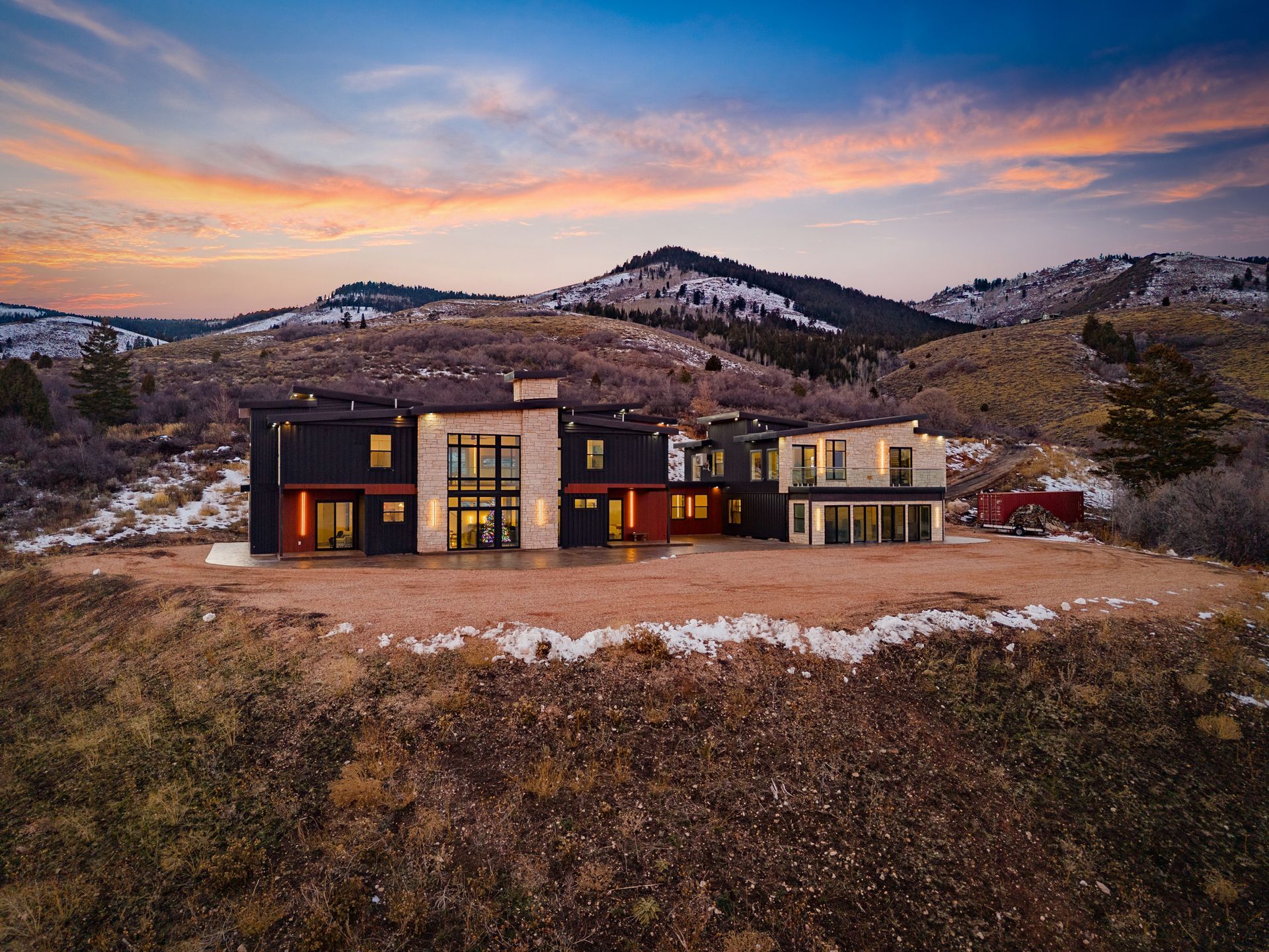 Modern hillside house at dusk with warm-lit windows, set against mountains and a pastel sunset