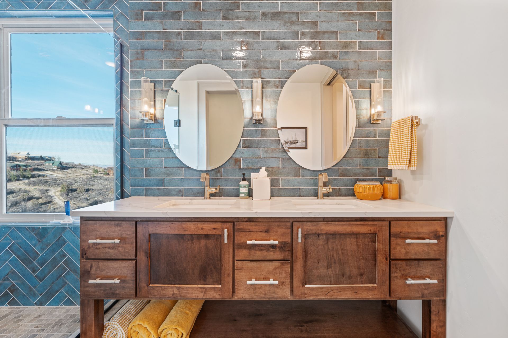 Bathroom vanity with two round mirrors, wood cabinets, white countertop, and blue-gray tile wall with window