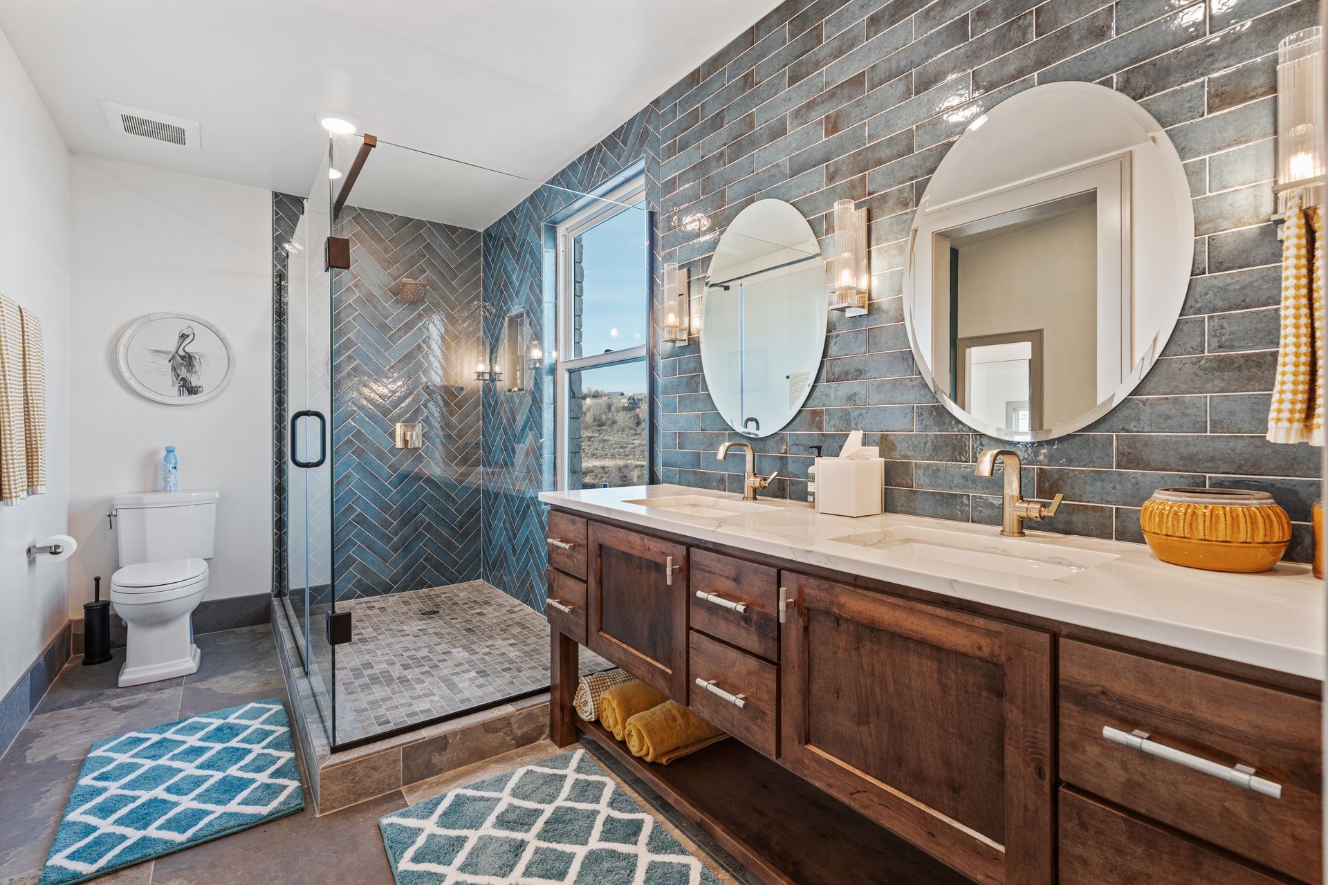Modern bathroom with double vanity, large mirrors, blue tile shower, and patterned floor mats