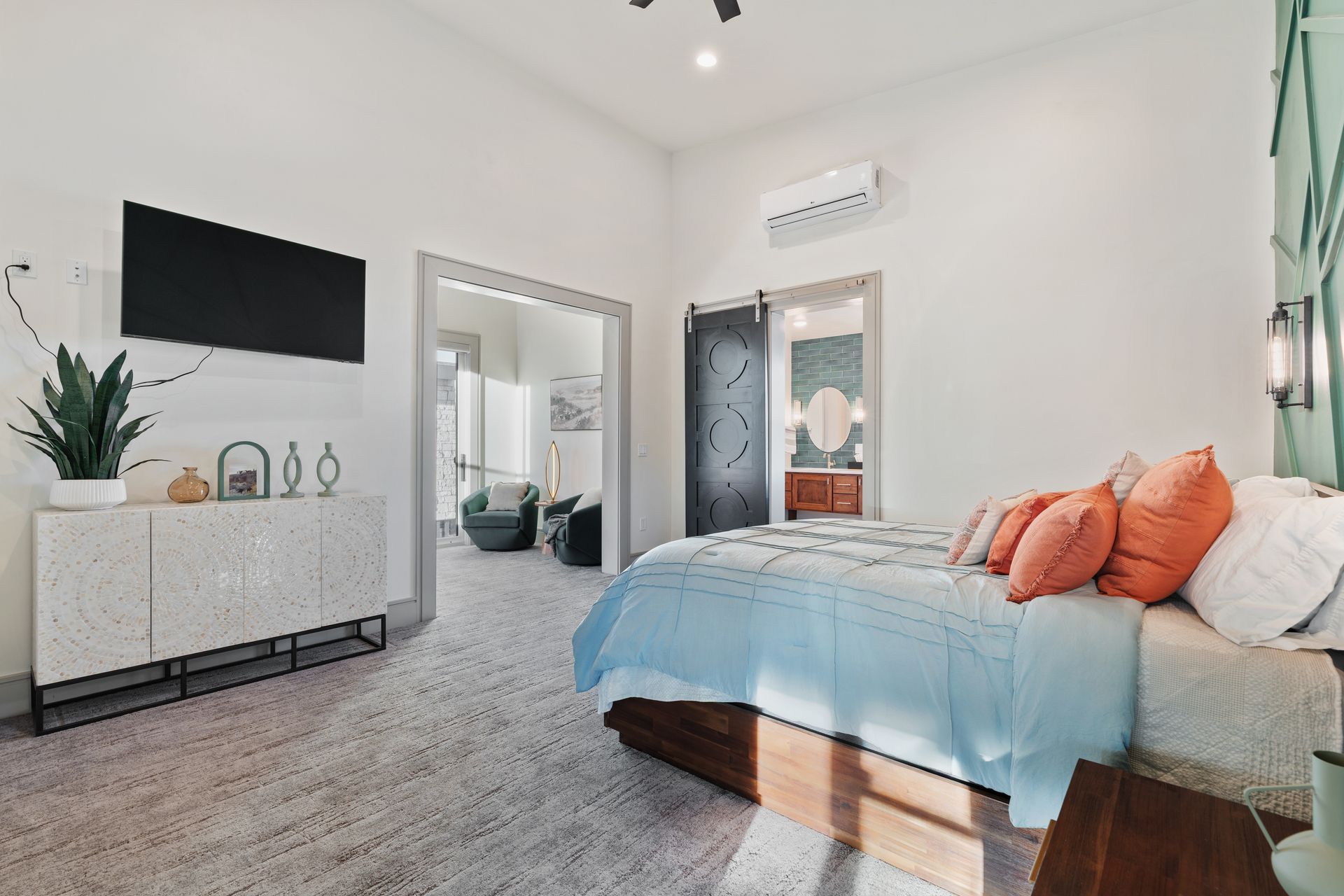 Bright bedroom with gray bedspread, coral pillows, wall-mounted TV, dresser, and mirrored closet doors