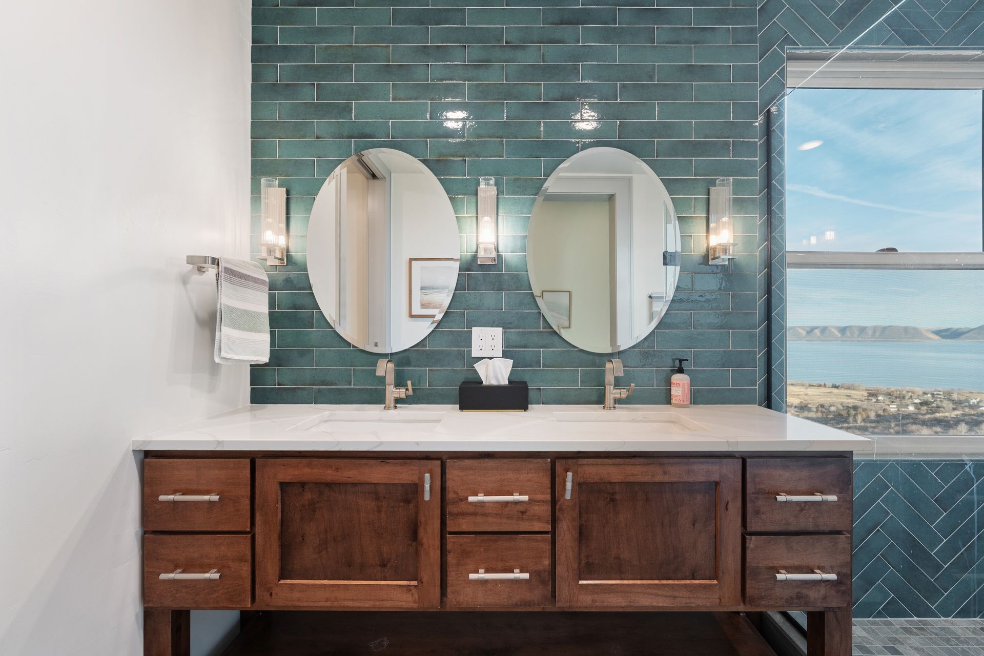 Modern bathroom vanity with two round mirrors, dark wood cabinets, and teal tile wall overlooking the ocean.