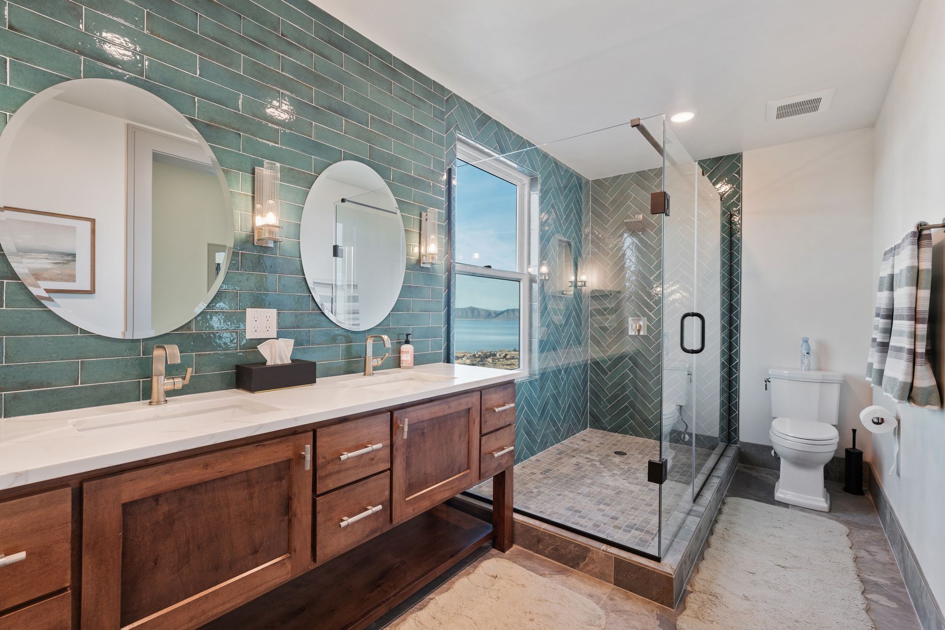 Modern bathroom with double vanity, teal tile walls, round mirrors, glass shower, and toilet