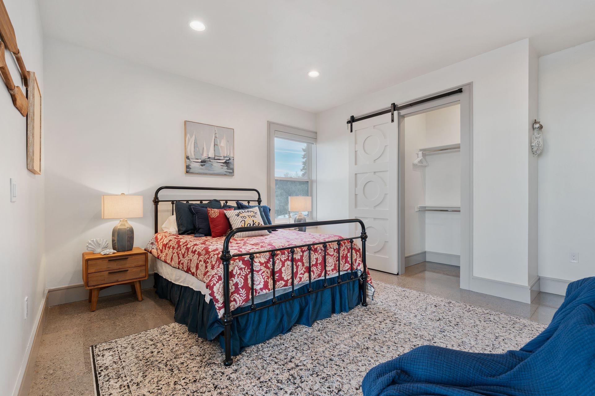Bright bedroom with floral bedspread, wooden nightstand, open closet, and blue chair on speckled floor.