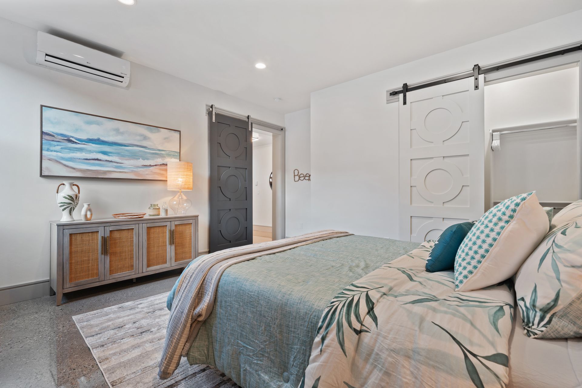 Bright bedroom with patterned bed, white walls, sliding barn door, and wicker console beneath beach wall art