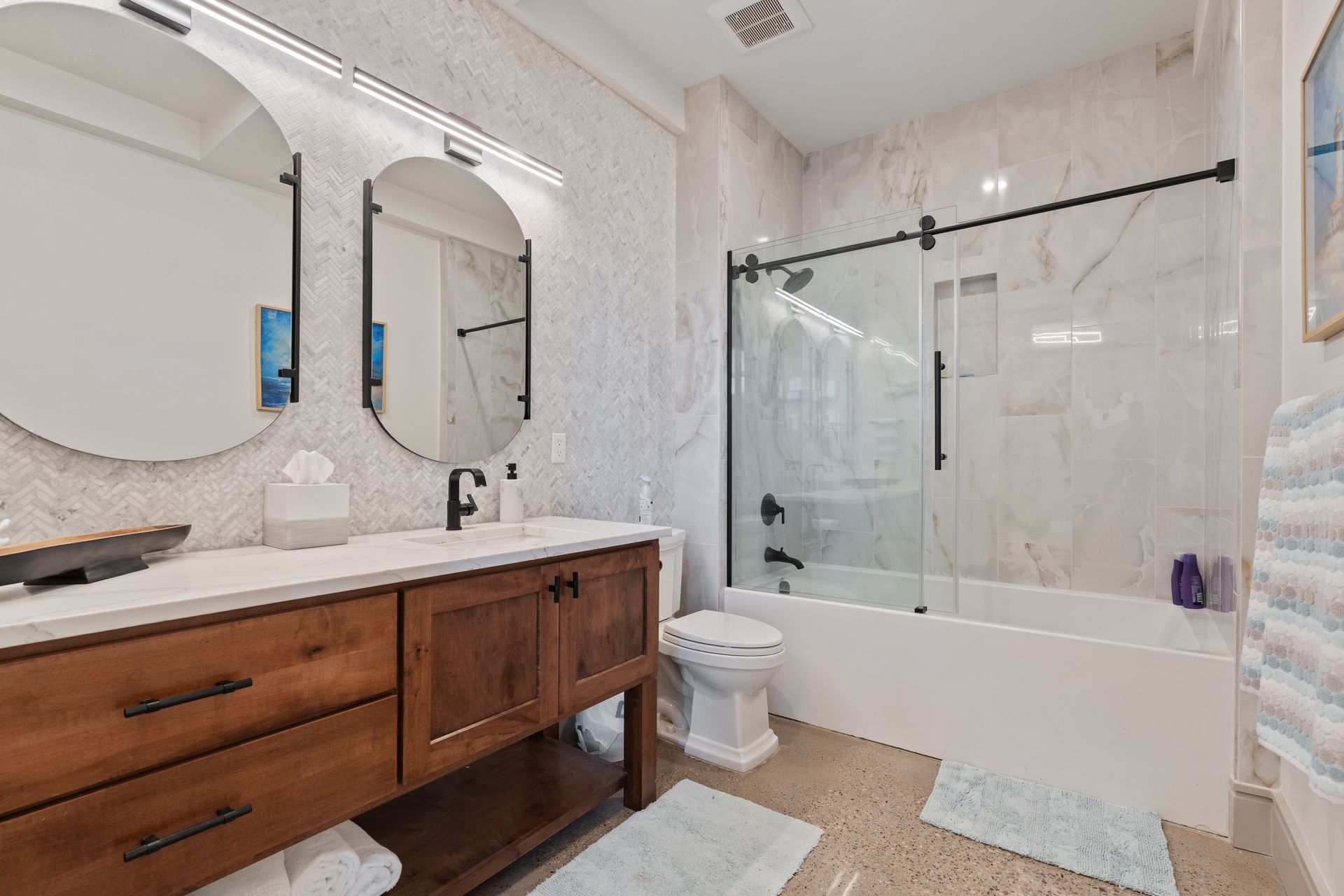 Bright bathroom with wood vanity, double mirrors, toilet, and glass shower over marble tile floor
