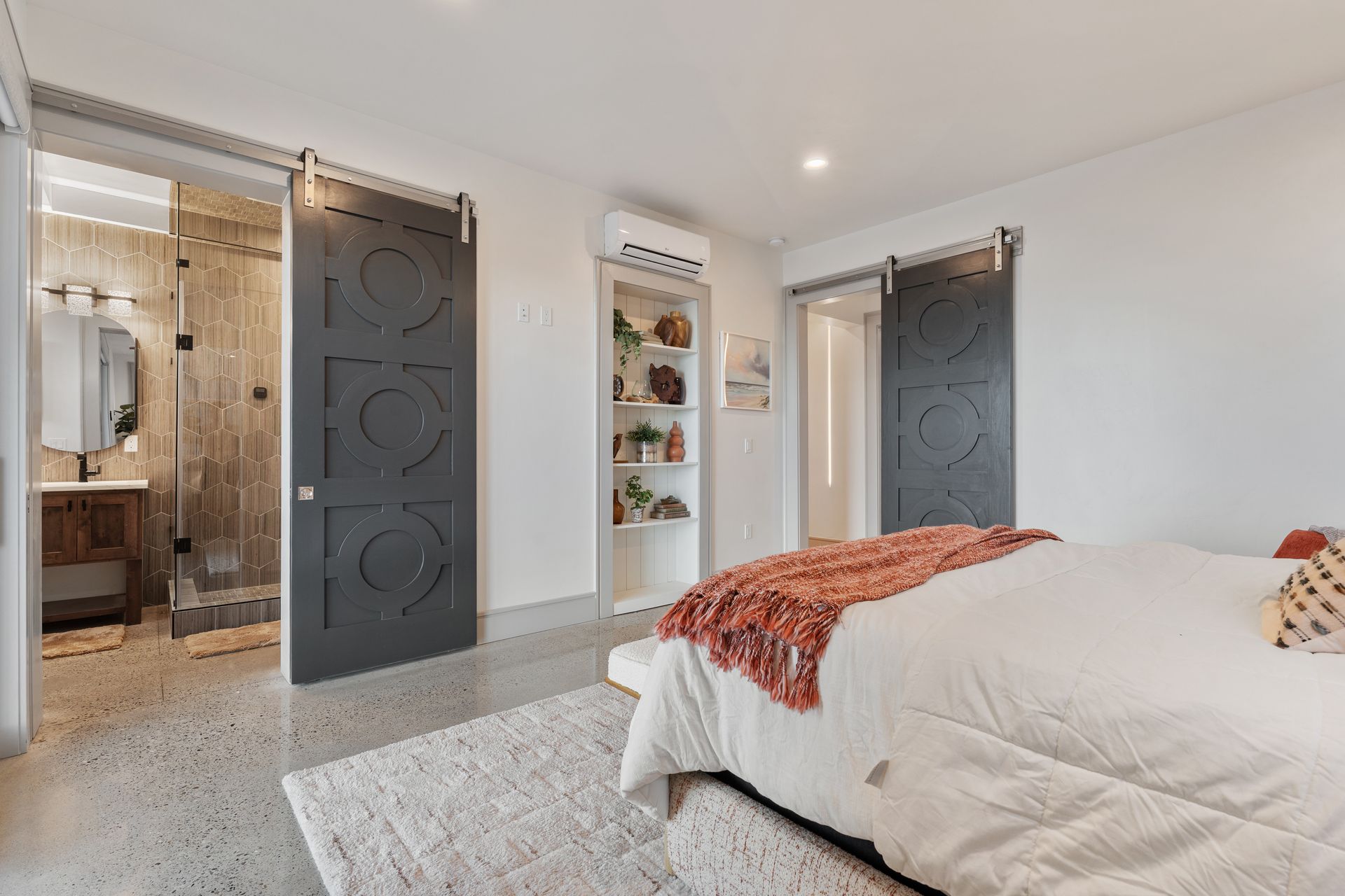 Modern bedroom with gray barn doors, white bedding, and an ensuite bathroom visible through an open doorway