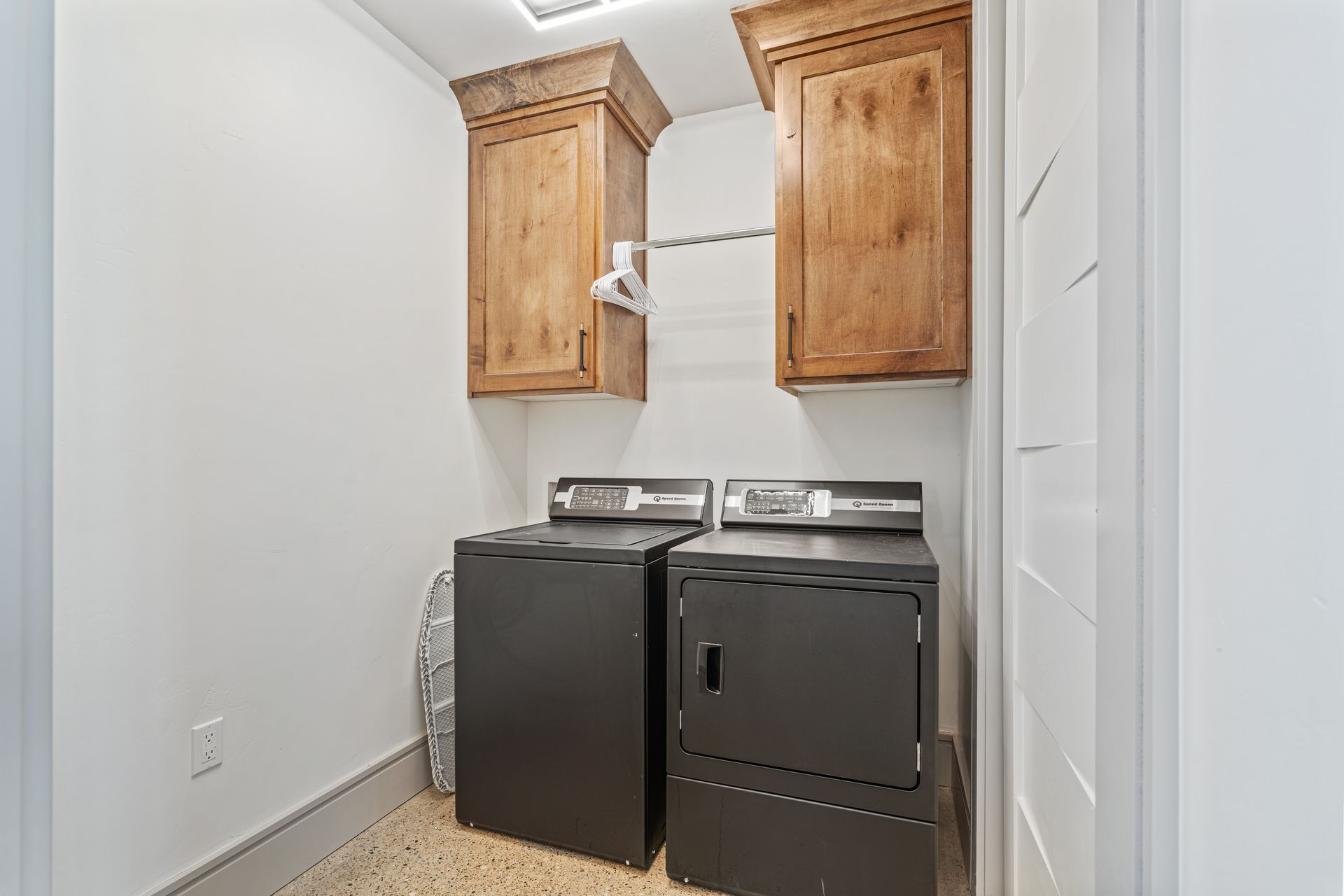 Laundry nook with a dark washer and dryer under wooden cabinets in a narrow white room.