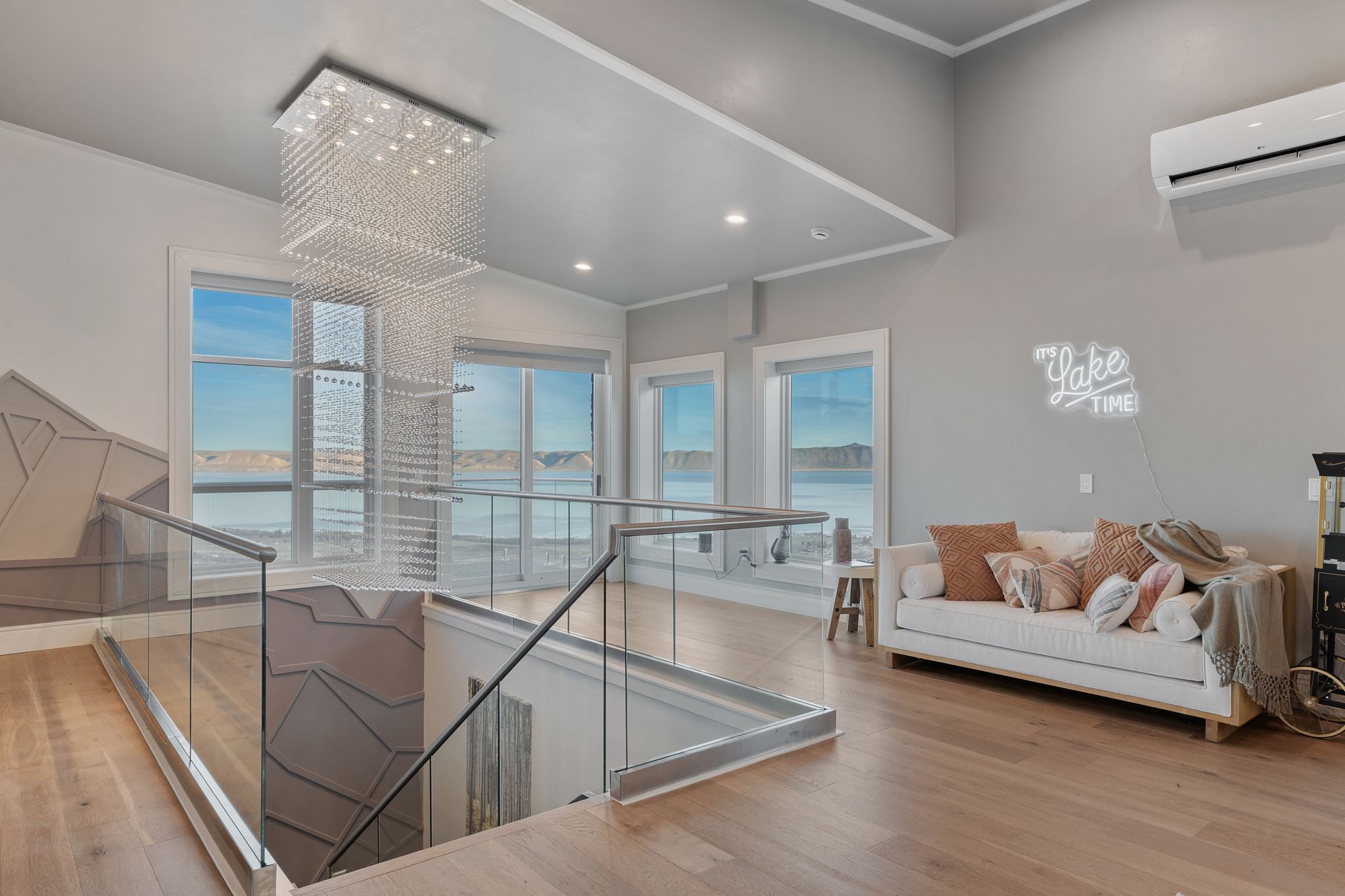 Modern living room with glass staircase, crystal chandelier, white sofa, and large windows with city view