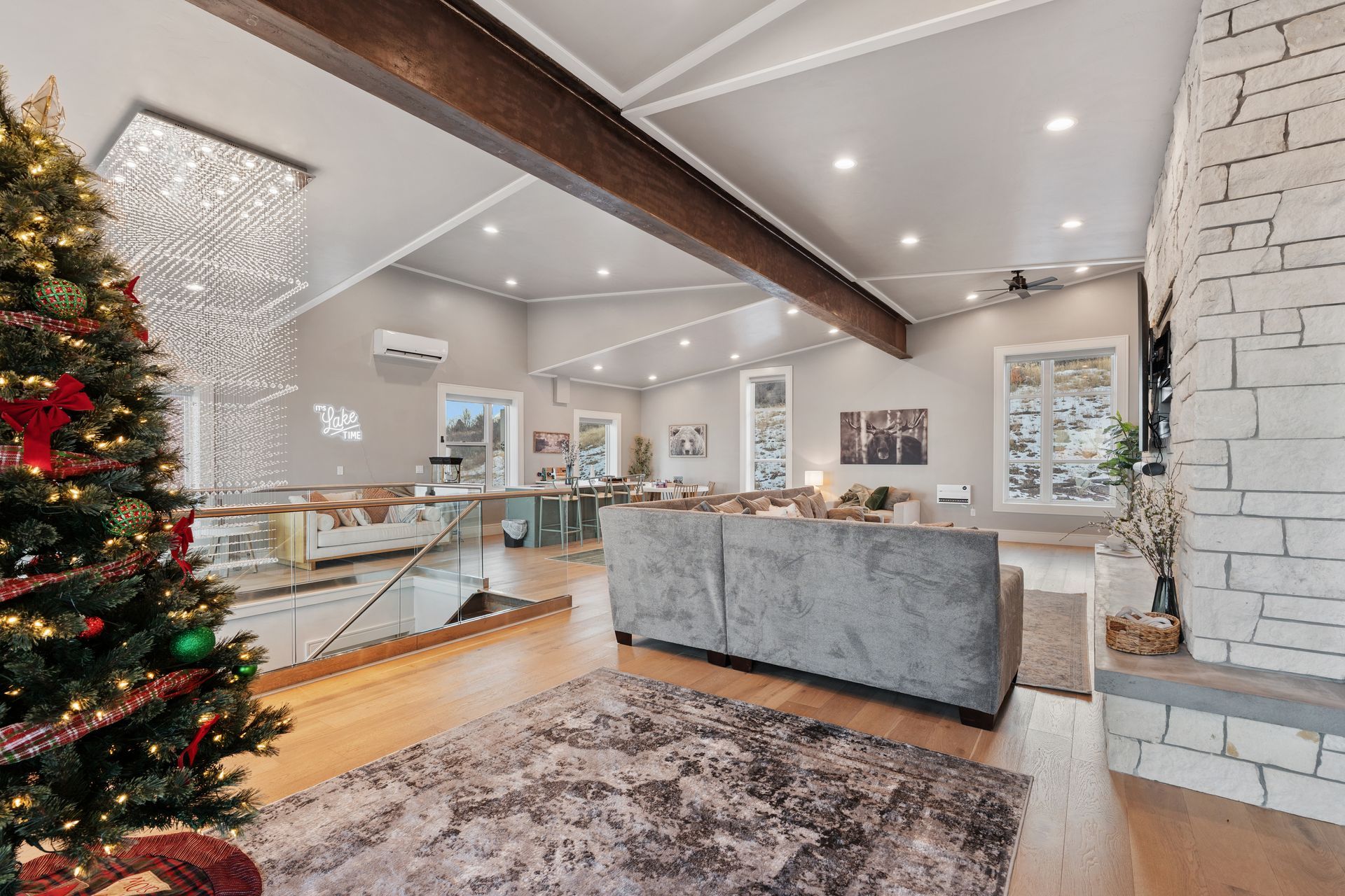 Bright living room with a Christmas tree, stone fireplace, gray sofa, and open kitchen area