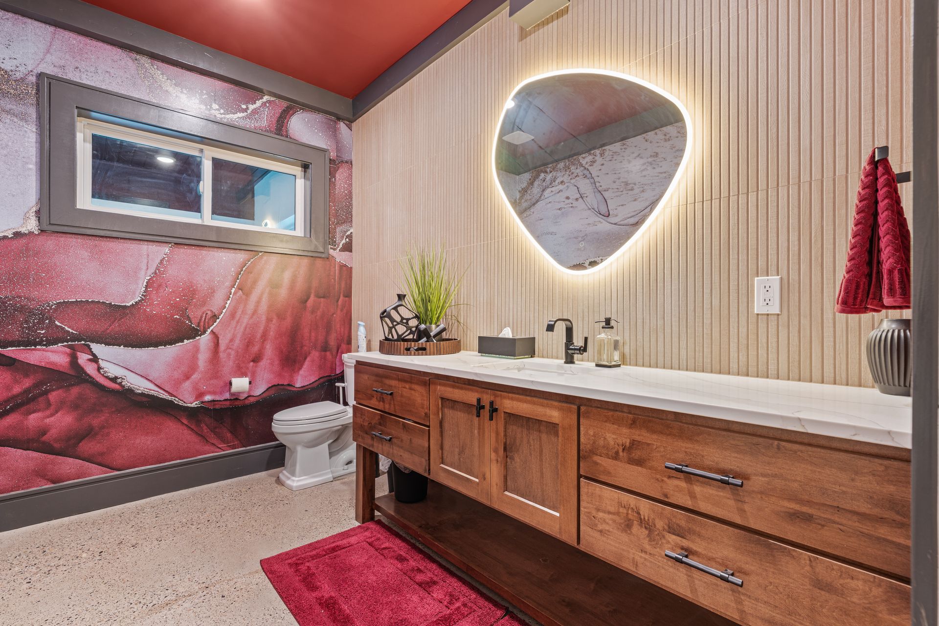Modern bathroom with wood vanity, lit round mirror, pink floral mural, and red accents