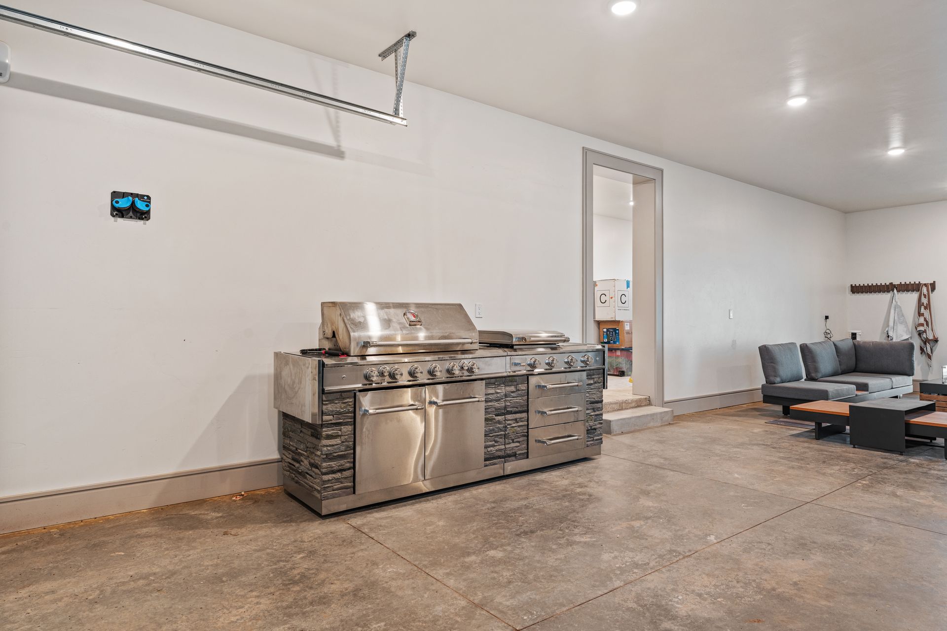 Spacious garage with stainless steel grill island, concrete floor, and gray sectional seating area.