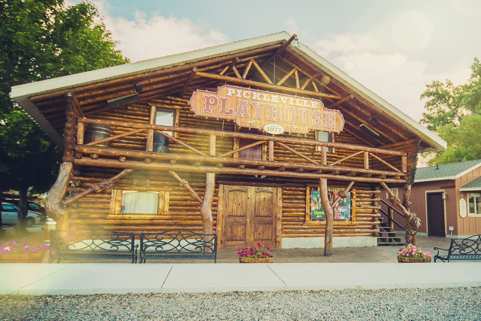 Rustic log cabin with a large sign, lit windows, and a gravel forecourt under a bright sky