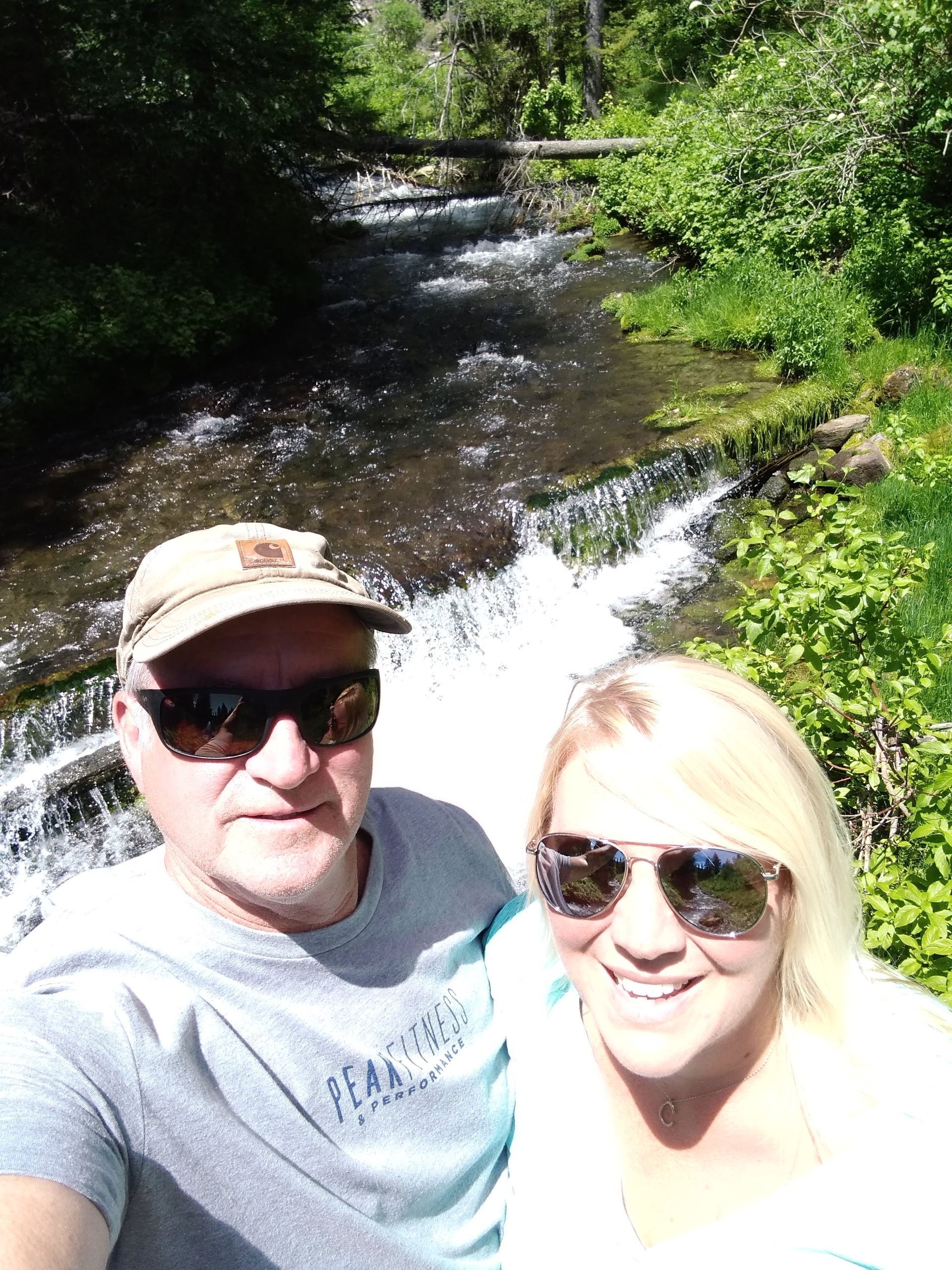 Two people smiling for a selfie by a sunlit forest stream and small waterfall