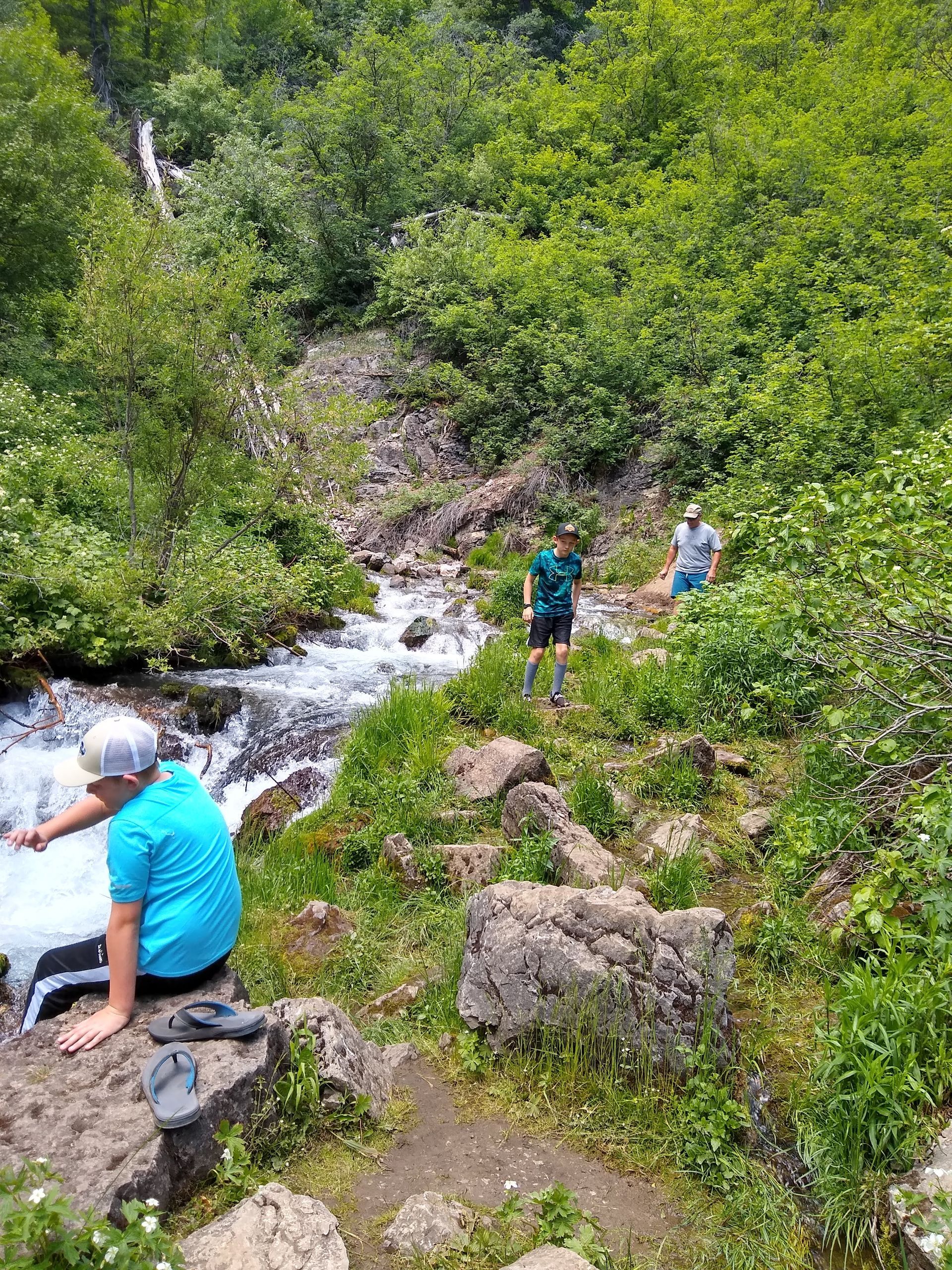 People resting beside a rushing stream in a lush green mountain trail area
