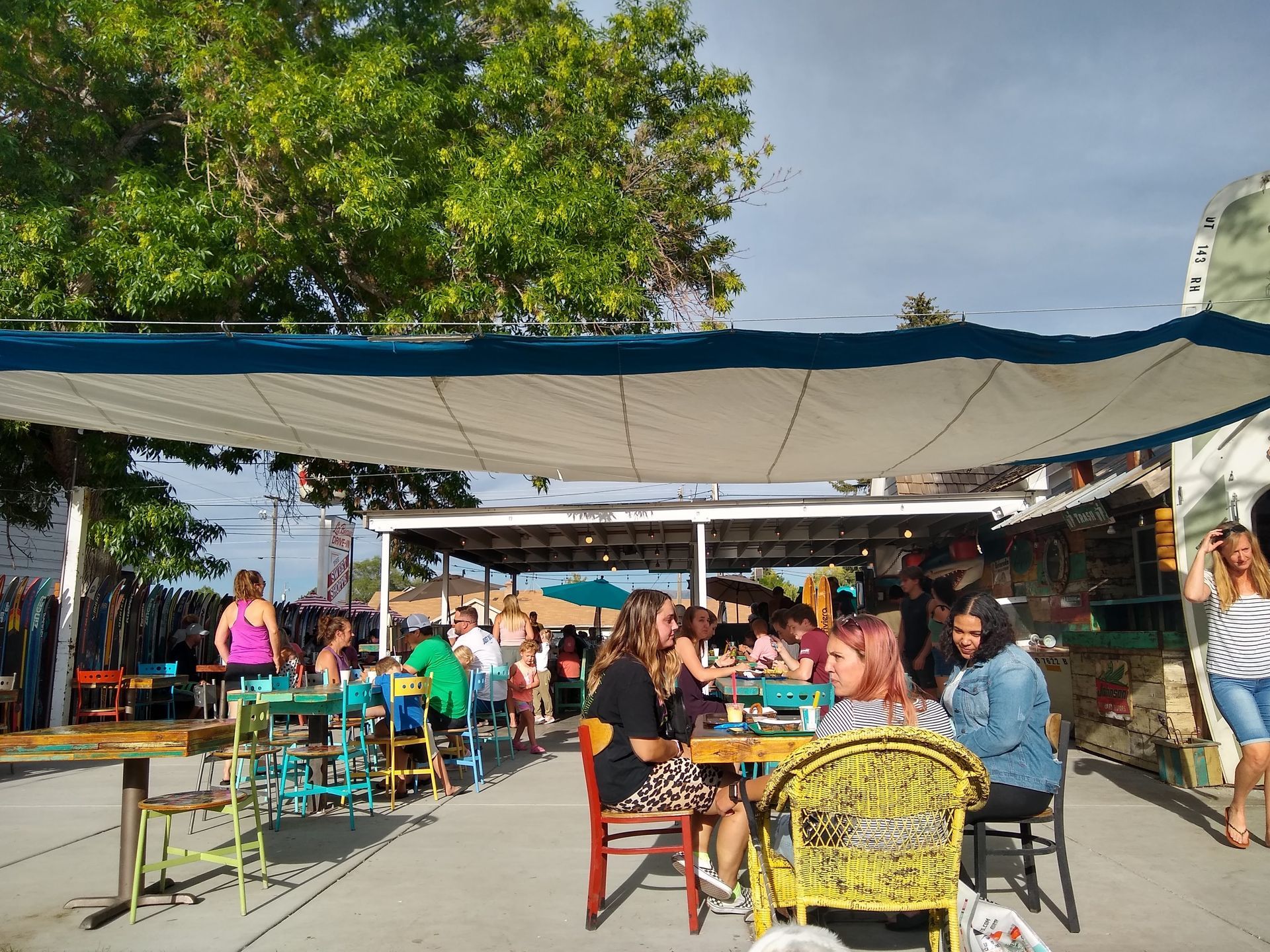 Outdoor café seating under a shade canopy with people eating and talking at colorful tables