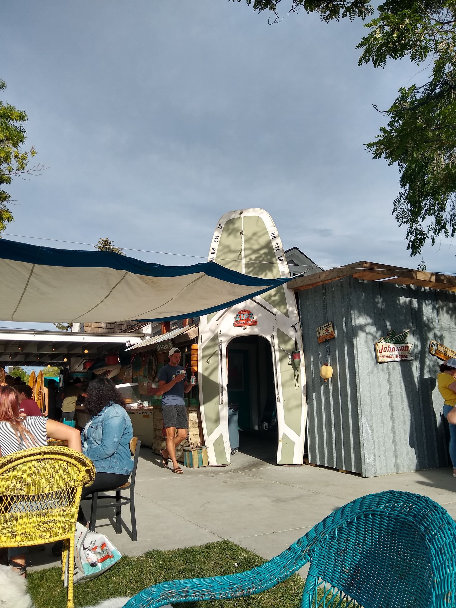 Outdoor café with shaded seating, a small white building, and people dining under trees.