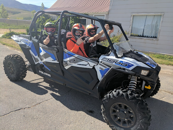 Three people in helmets riding a blue and black side-by-side off-road vehicle on a paved road.
