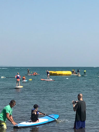 People paddle and float in a calm blue sea near a large yellow inflatable obstacle.