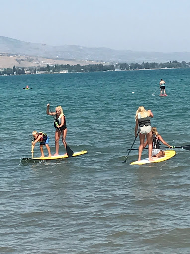 People paddleboarding on a lake, with swimmers and distant shoreline in the background.